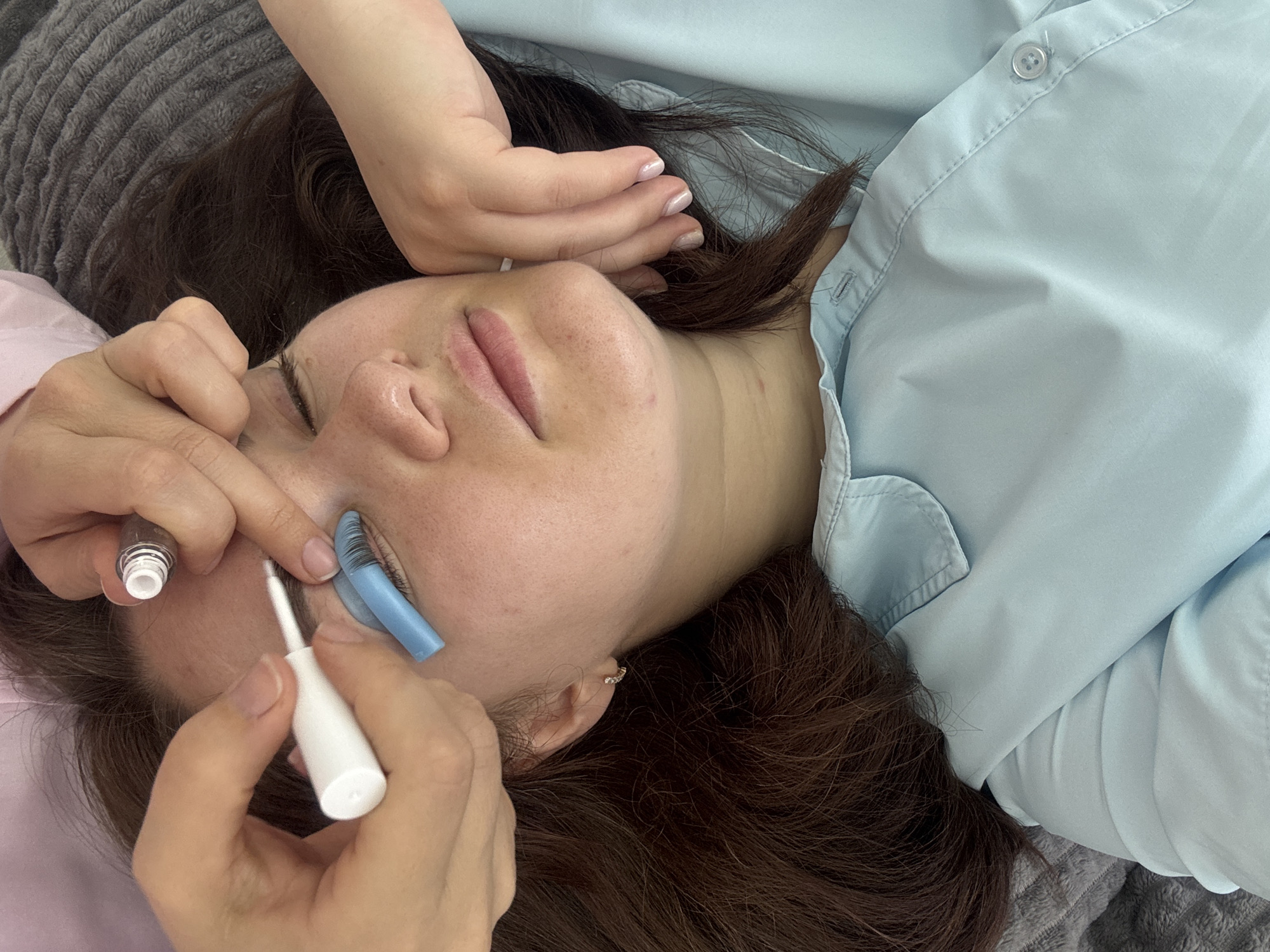 This is a top-down, close-up shot of a woman lying down, undergoing an eyelash treatment. The woman has her eyes closed and is wearing a light blue button-down shirt. Her dark brown hair is spread out on a grey textured surface, likely a blanket or couch. Two pairs of hands are visible, one applying a white substance to the woman's eyelashes with a brush, the other holding a small clear bottle with a white cap. A blue silicone pad is positioned under the woman's left eyelid. The scene appears to be indoors, but no specific city location is discernible from the visual information.
