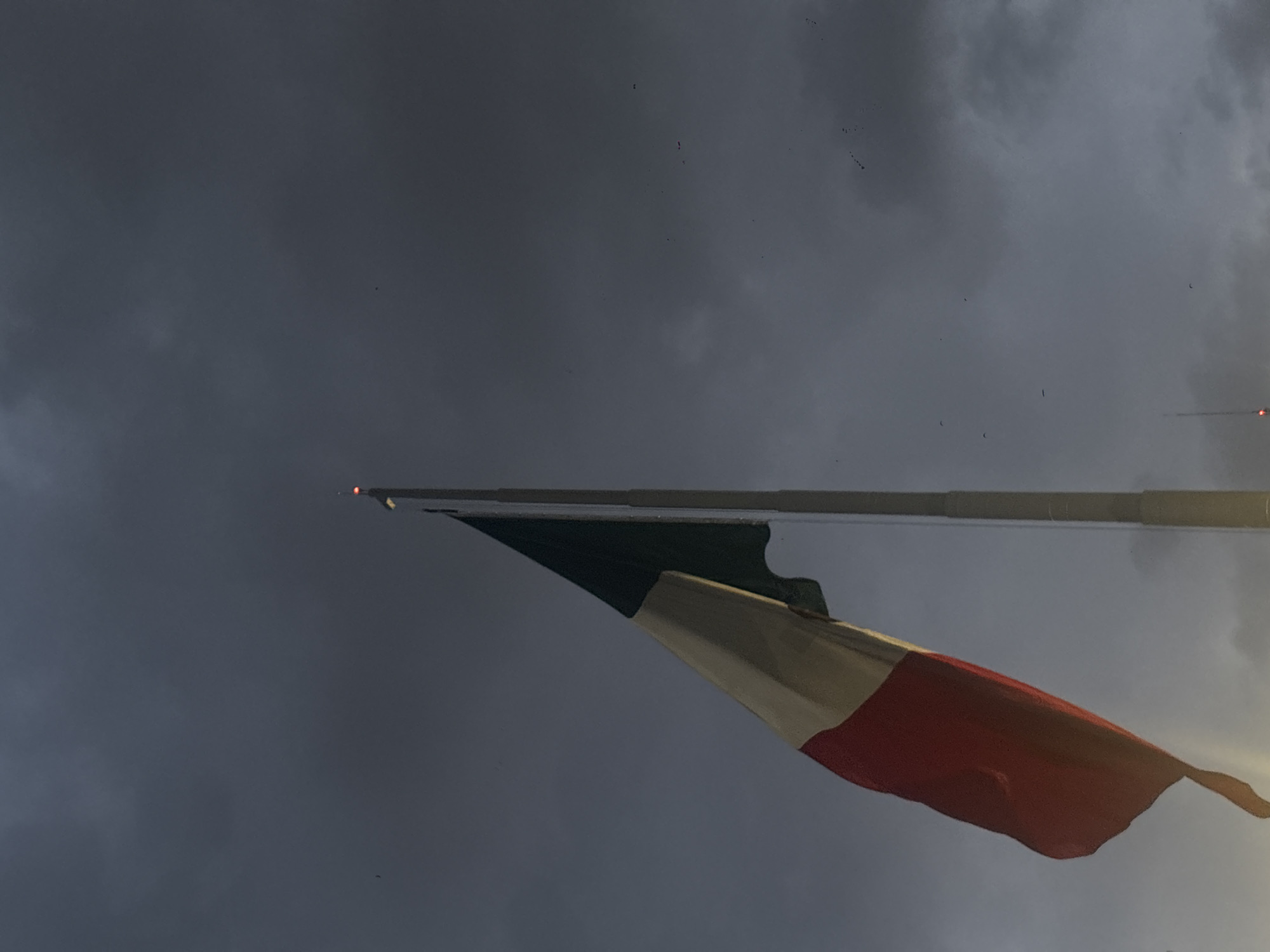 The image shows a French flag blowing in the wind against a cloudy sky. The flag is mounted on a pole, and the colors of the flag (blue, white, and red) are visible. The image was likely taken in Mexico City, Mexico.
