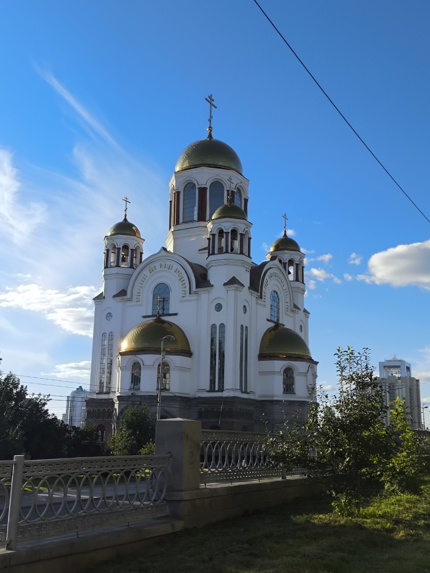 This is a full-shot, eye-level, outdoor, vertical photograph of the Church on Blood in Yekaterinburg, Russia. The Church on Blood is a Russian Orthodox church built on the site of the Ipatiev House, where Nicholas II, the last emperor of Russia, and his family were executed. The church is white with golden domes and crosses. It is surrounded by trees and a fence. In the background, modern buildings can be seen. The sky is blue with some clouds and a power line stretches across the top of the frame. The sun is shining, casting shadows and highlighting the golden domes. There are no people visible in the image.