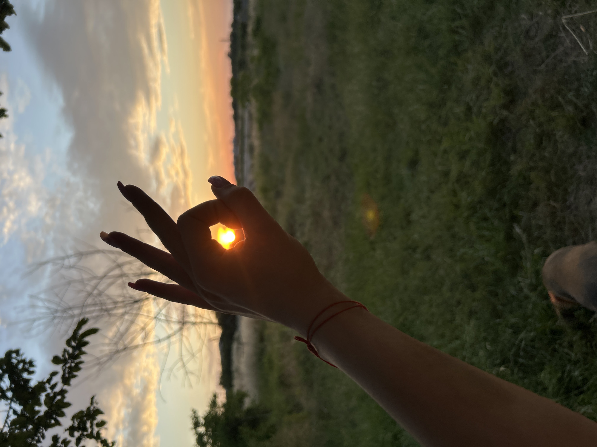 A hand forms an "okay" symbol, framing a setting sun visible through the gap between the thumb and forefinger. The sun, appearing as a bright orange orb, casts a warm glow on the skin. The hand has manicured fingernails painted a dark color, and a thin red bracelet is worn around the wrist. The background depicts a blurry landscape with green grass and distant trees. The sky is filled with soft clouds illuminated by the sunset, transitioning from pale blue to hues of orange and yellow. The overall scene evokes a sense of calm and tranquility, capturing a moment at dusk. The orientation of the image is vertical.
