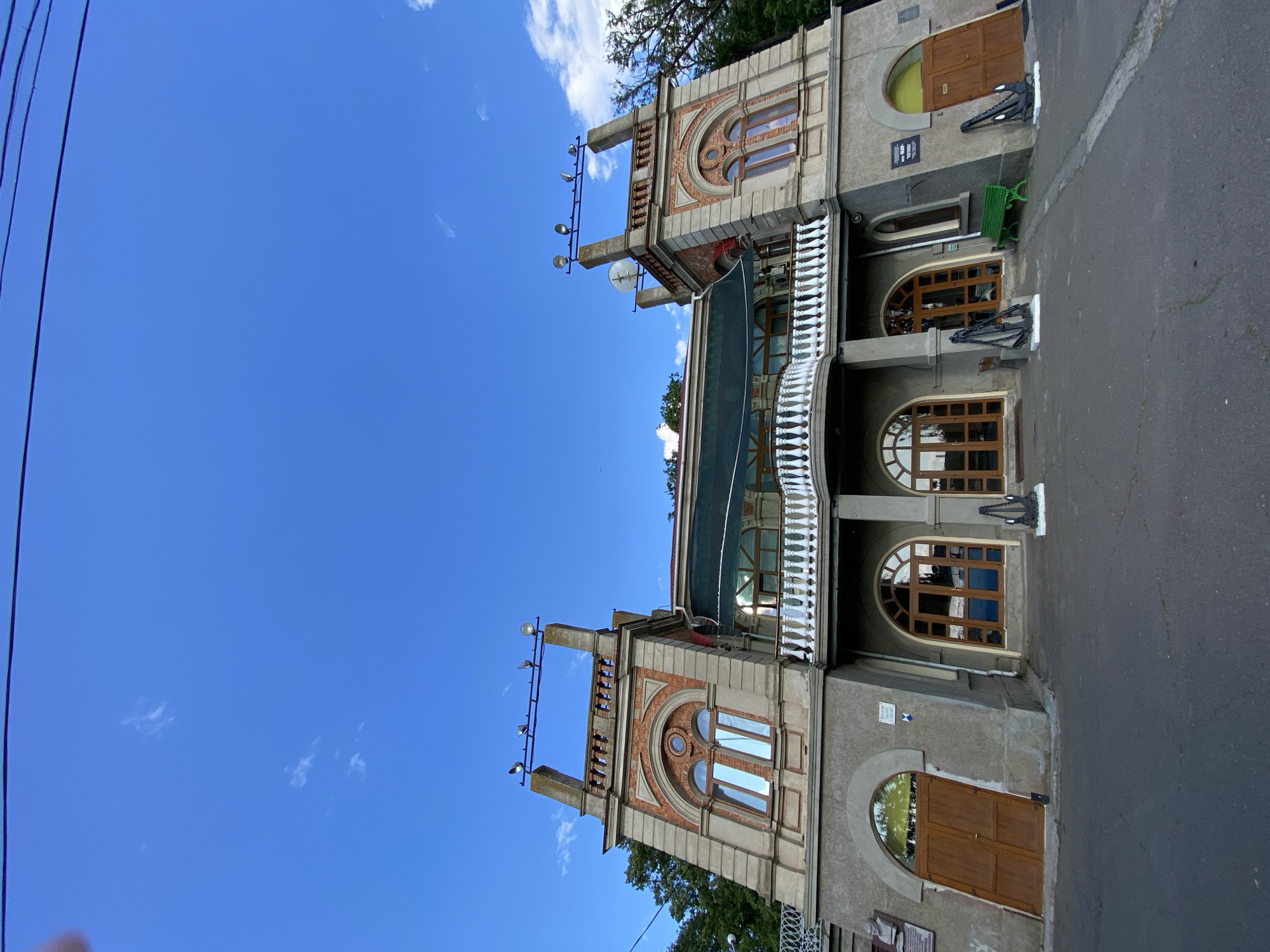 An exterior, low-angle, eye-level shot captures the facade of a two-story building with a symmetrical design. The building features a central section with a balcony and two flanking towers. The towers have arched windows and are topped with decorative railings and what appear to be spotlights on poles. The main facade is constructed of concrete, with brick accents around the upper windows and decorative elements. Arched doorways with wooden doors are visible on the ground floor. Several decorative anchors are placed on pedestals outside the building, and a green bench is situated near the right side. The scene is set against a bright blue sky with a few wispy clouds and some trees are visible on the left and right sides. Power lines are visible crossing the top of the frame.