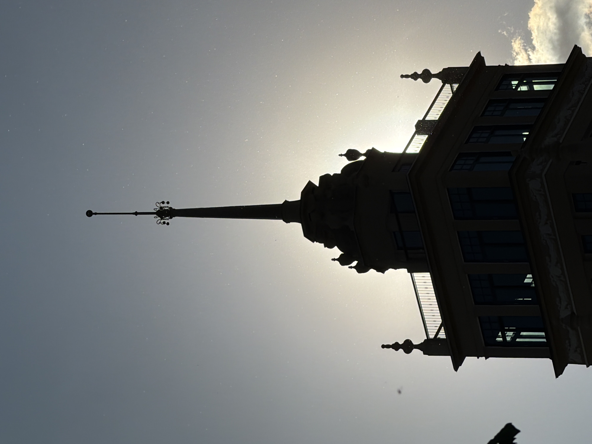 The image is a low-angle, silhouetted view of a building's spire and upper floors against a bright, hazy sky. The building's architecture features a tall, pointed spire with decorative elements at the top and along its length. The structure also includes visible windows and a balcony. The lighting suggests that the sun is behind the building, causing it to appear as a dark silhouette. The image is oriented in a portrait format, with the building appearing to be positioned sideways. Based on the architectural style and spire, this could potentially be a building in Moscow, Russia, due to similar structures present in the city.