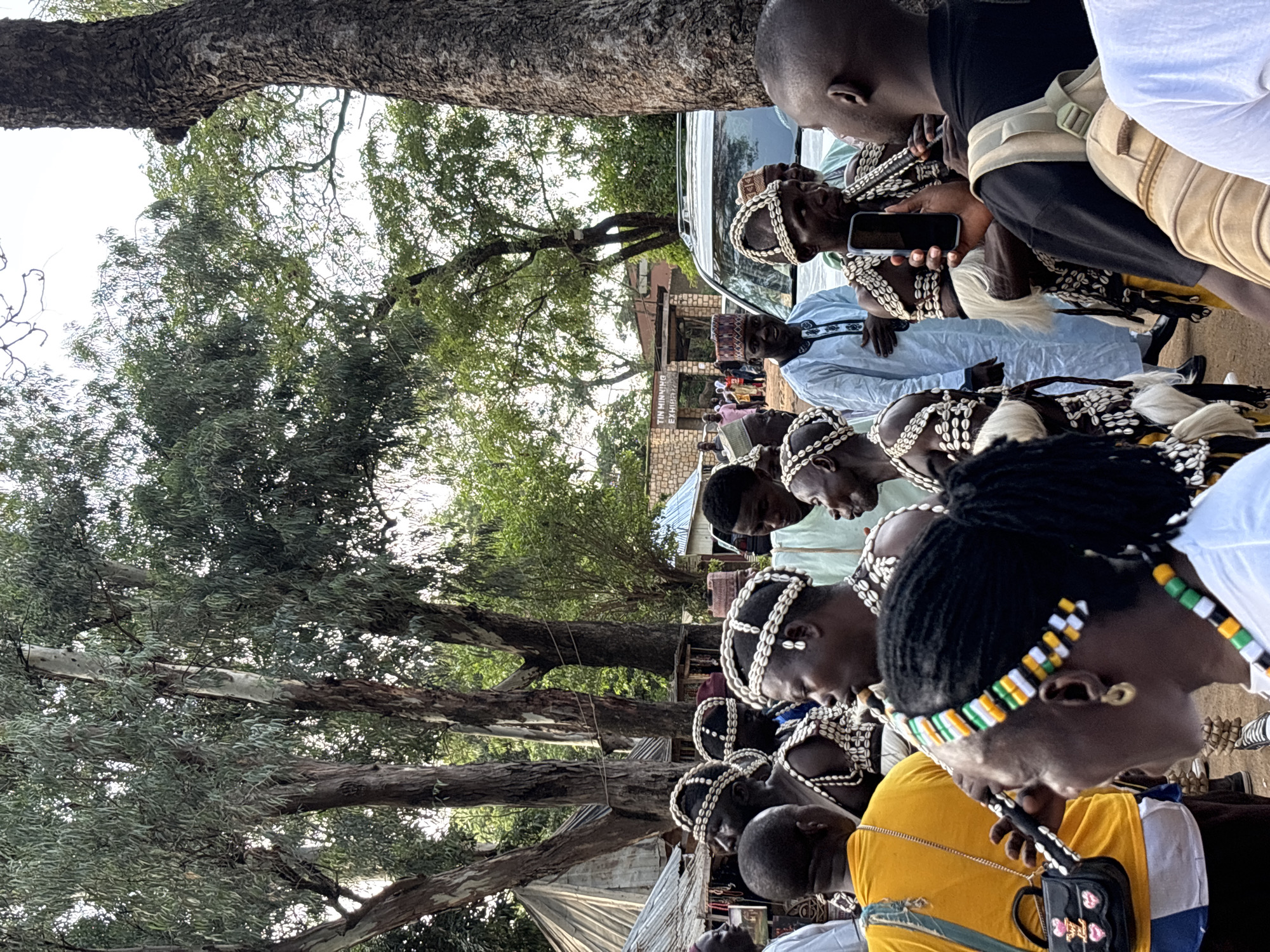 The image depicts an outdoor gathering in Jos, Nigeria. In the foreground, a group of people are visible, some adorned in traditional attire featuring shell necklaces and headdresses. A person in a bright yellow shirt is holding a small black handbag. To their right, a woman with a beaded headband and necklace is seen in profile. In the midground, several men wearing elaborate shell decorations and white skirts are standing, with one man playing a flute-like instrument and holding a mobile phone. Another man in a light blue robe and a patterned cap is looking towards the camera. Behind them, a white SUV is parked, and a building with text reading "TIN MINNING EXHIB". Several trees with large trunks and green foliage dominate the background, and glimpses of tents and other structures suggest a market or event setting.