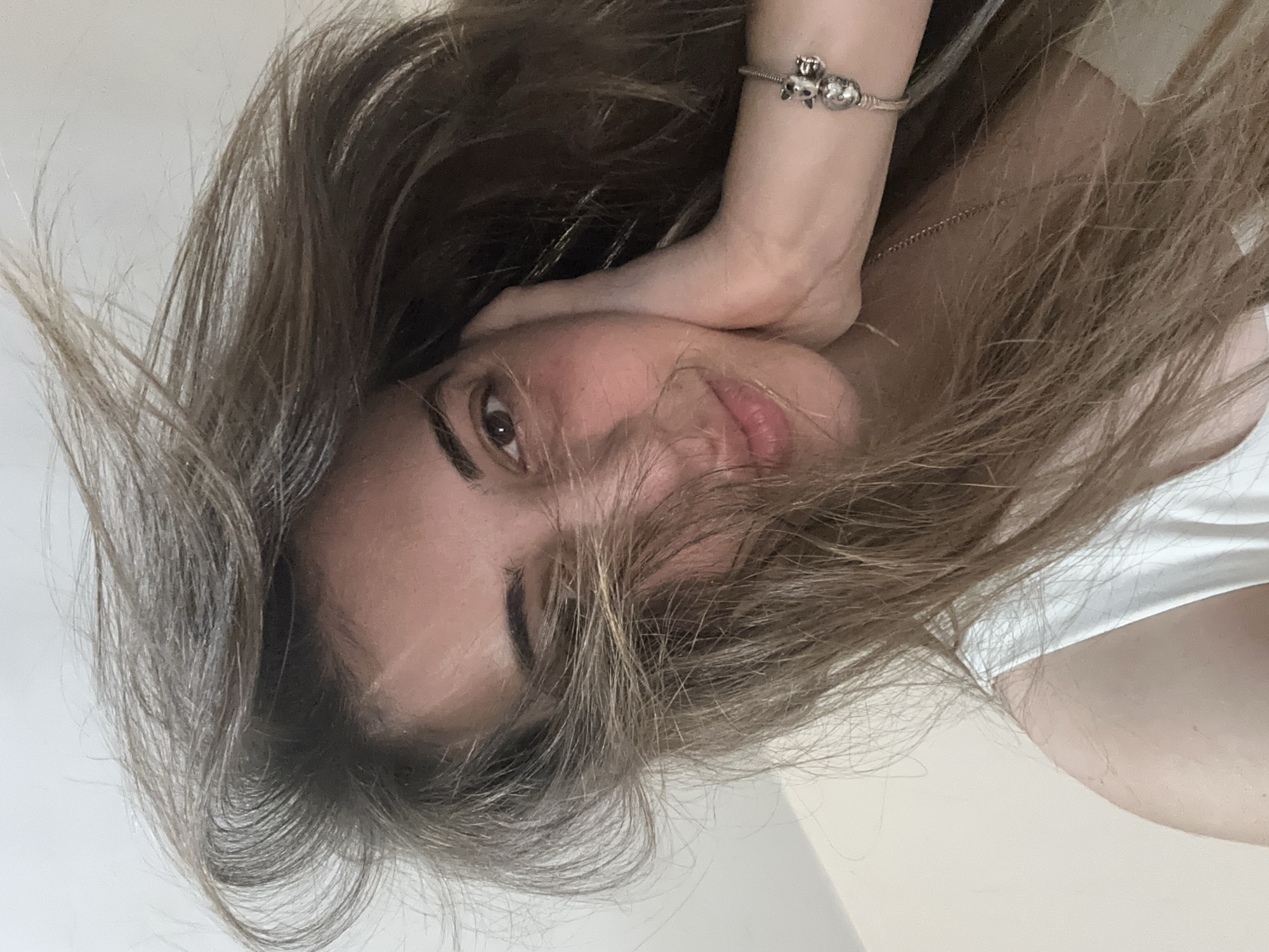 The image is a close-up, tilted view of a woman with long brown hair. She is resting her face on her hand, which has a silver bracelet with charms. She is wearing a white top. The background is a plain, light-colored wall. No location or context suggests the scene is in Sirakovo, Bulgaria.