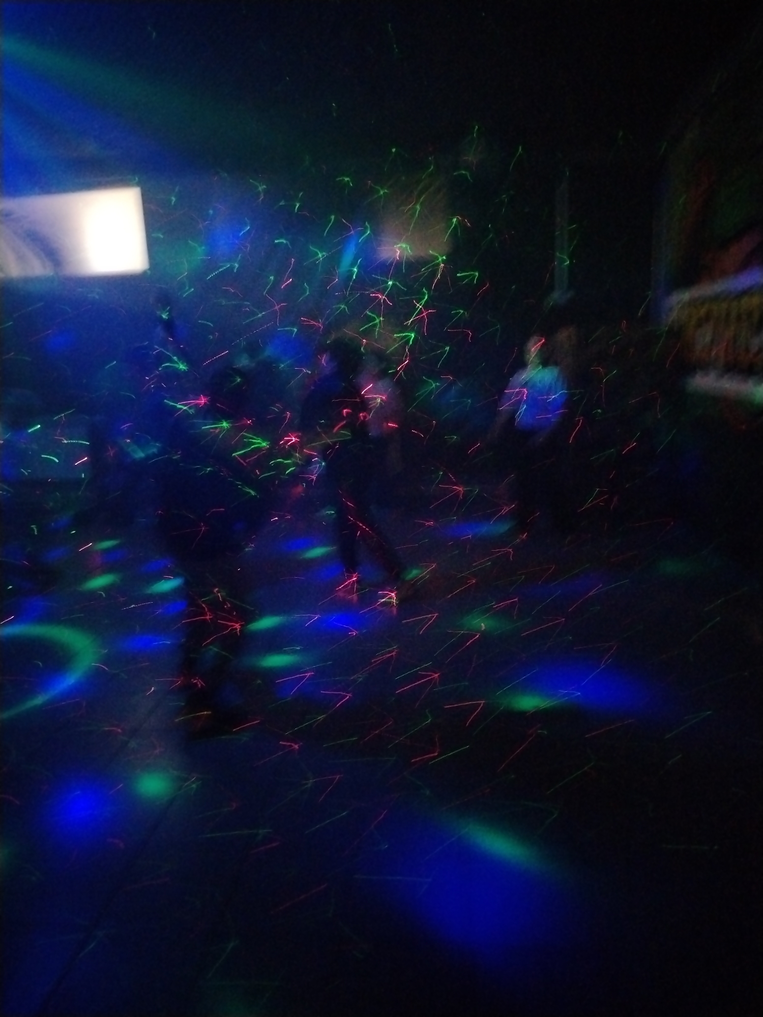 The image appears to be taken inside a dimly lit room, potentially a nightclub or event venue. Several indistinct human figures are present, suggesting a gathering or social event. The environment is illuminated by a dynamic array of green, blue, and red laser lights that create streaks and patterns throughout the space, including across the floor. The location in Rusanovo, Russia is not explicitly verified by the image content and is based on the user-provided context.