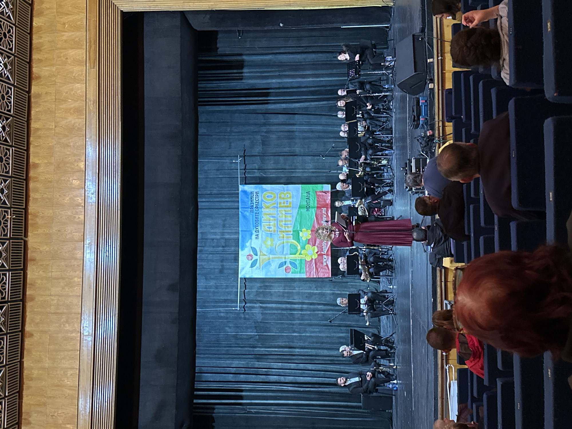 The image captures a performance in Montana, Bulgaria. On stage, an orchestra is seated, with musicians in black suits and music stands in front of them. A banner hangs behind the musicians, displaying the words "Днико Ниоиев". A woman in a maroon dress stands in front of the orchestra, seemingly a performer or singer. In the foreground, audience members are visible in theater seating. The environment appears to be a theater or auditorium, with wood-paneled walls on the left side of the image and dark curtains behind the stage.