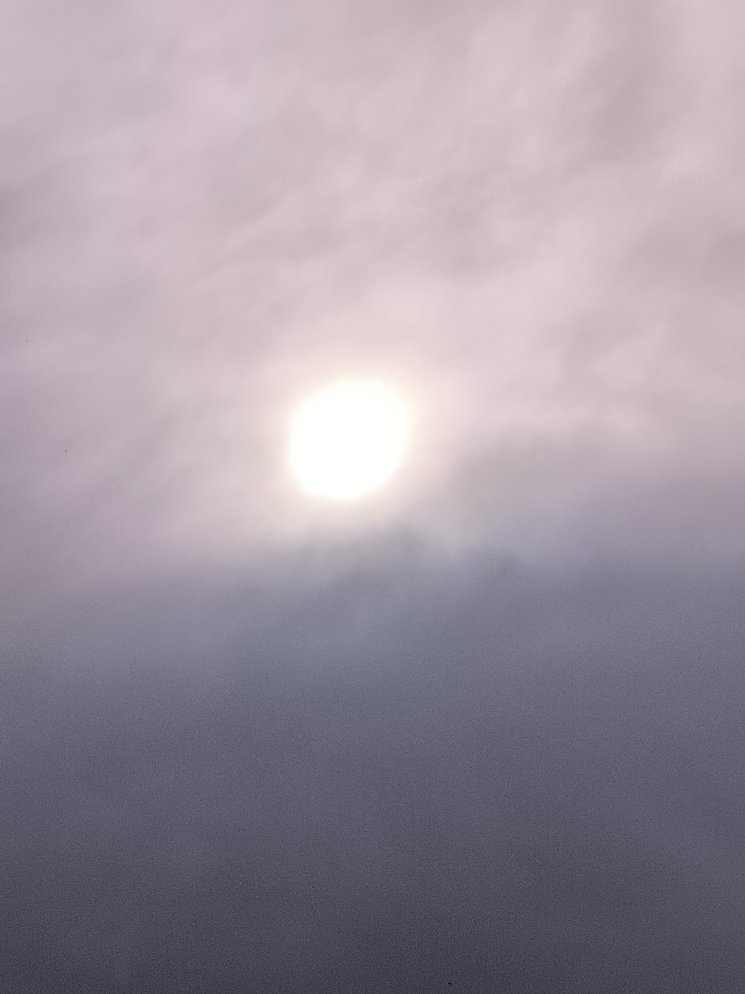 This media file is a vertical photograph taken outdoors, featuring a hazy sky with a bright, diffused sun prominently positioned slightly above the center. The sun appears as a luminous, almost white orb, its brightness creating a soft glow that permeates the surrounding atmosphere. The sky is filled with wispy, light-colored clouds that are tinged with a pale purple or pink hue, suggesting either a sunrise or sunset. The lower portion of the image is dominated by darker, denser clouds that create a gradient effect, transitioning from the lighter sky above to a deep, muted gray-purple at the bottom. There are no discernible people, objects, or man-made structures in the frame. The overall scene evokes a sense of atmospheric diffusion and muted light. The time of day is likely dawn or dusk, given the quality of light and cloud colors. The weather appears overcast or foggy, with the sun's rays softened by the atmospheric conditions. No text is visible in the image.