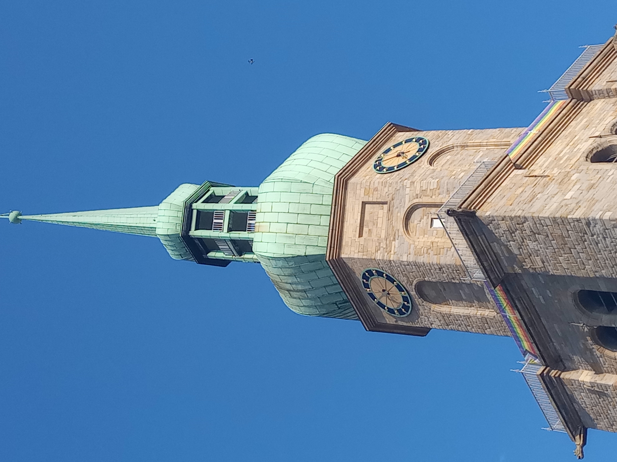 The image shows the tower of the Reinoldikirche in Dortmund, Germany, against a clear blue sky. The tower is constructed of light-colored stone blocks. It features two circular clocks with dark blue faces and yellow hands. A rainbow-colored banner is draped over a railing on one side of the tower. The tower is topped with a green, multi-tiered spire. A small dark object, possibly a bird, is visible in the sky to the right of the spire. A gargoyle-like carving is visible on the lower left side of the tower.