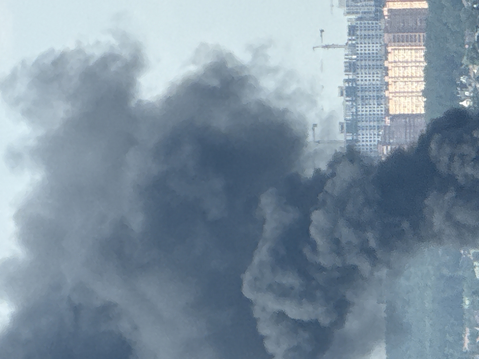 A plume of dark smoke billows into the sky, obscuring the upper portion of the frame. In the middle ground, a densely packed urban landscape unfolds, characterized by rows of apartment buildings of varying heights and architectural styles. Some buildings are clad in light-colored materials, while others feature orange facades. Cranes are visible amidst the buildings, suggesting ongoing construction. In the foreground, a line of trees partially conceals a smattering of smaller buildings, indicating a residential area bordering the city. The overall scene suggests a post-industrial or developing urban environment, with the dominant element being the large smoke plume, hinting at a potential fire or industrial event. The image captures a wide view of the city of Lyubertsy, Russia, under a hazy sky.
