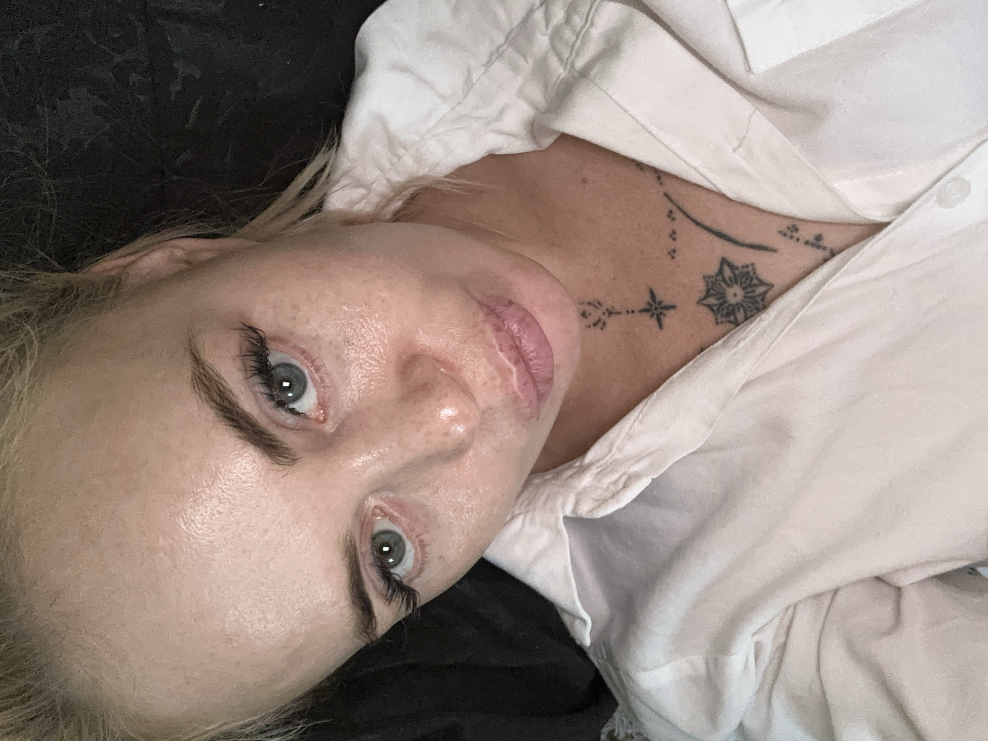 The image is a close-up, high-angle, selfie-style portrait of a fair-skinned woman with blonde hair tied back. She is wearing a white collared shirt, buttoned at the collar. Her eyes are open and looking directly at the camera, with long, dark eyelashes. She has light eyebrows and a fair complexion with some freckles visible. Her lips are slightly parted and appear to have a pinkish hue. A tattoo is visible on her chest, featuring geometric patterns and lines. The background is dark and out of focus. The lighting appears to be overhead and slightly to the side, highlighting the sheen on her skin. The scene is indoors and the location is unknown.