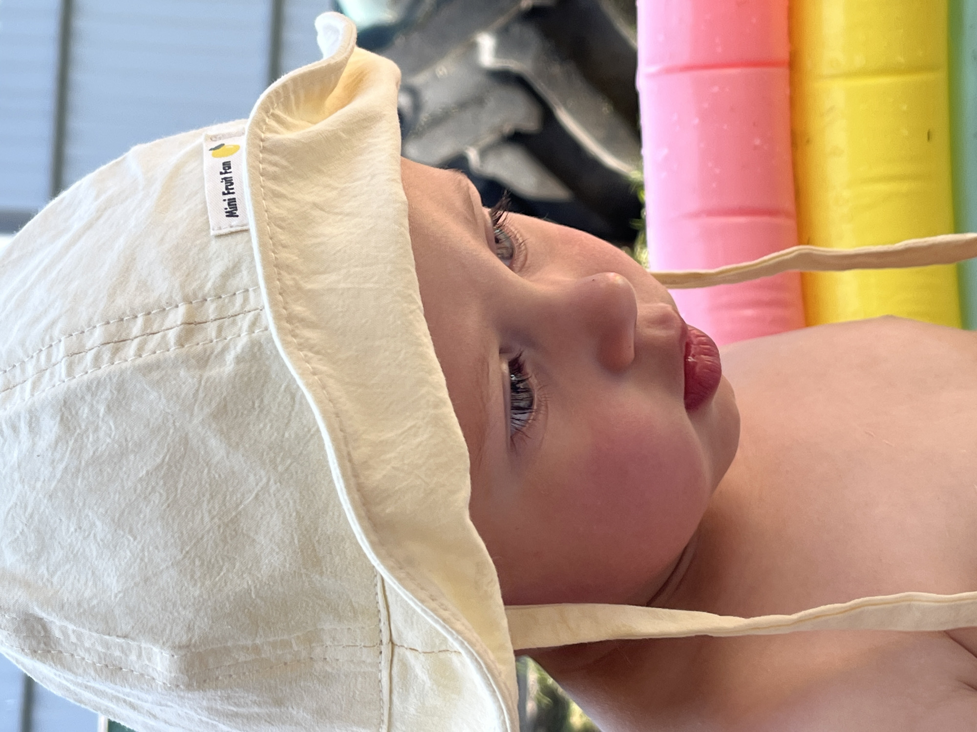 A close-up shot depicts a baby lying down, wearing a cream-colored sun hat with a tag labeled "Mini Fruit Fun." The baby's face is visible in profile, showing their nose, lips, and one eye. The background includes blurred objects in various colors, including pink and yellow. The location Фоки, Russia is not discernible from the visual information available in the image.