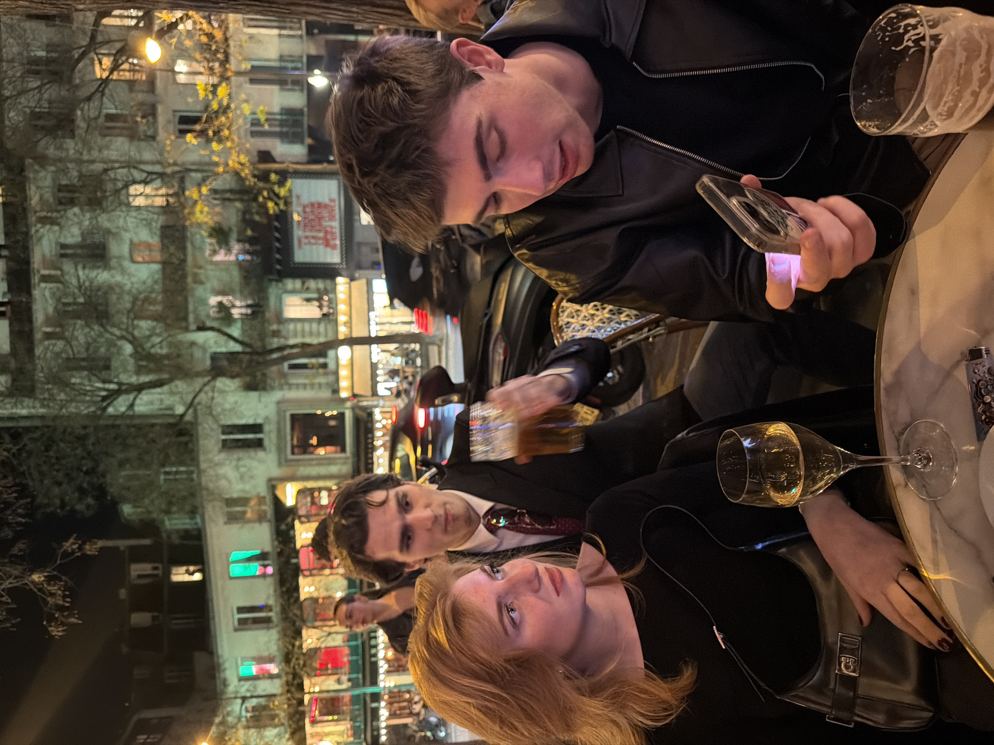 Three individuals are at an outdoor table, likely at a restaurant or café. One person, wearing a black leather jacket, is holding a smartphone and appears to be using it. Another person is in a suit and tie. A woman with reddish-blonde hair is visible. The table has various drinks, including a glass of beer and a wine glass. Buildings and street lights are in the background, suggesting an urban setting. The location seems to be in Paris, France, with the presence of Parisian-style buildings.