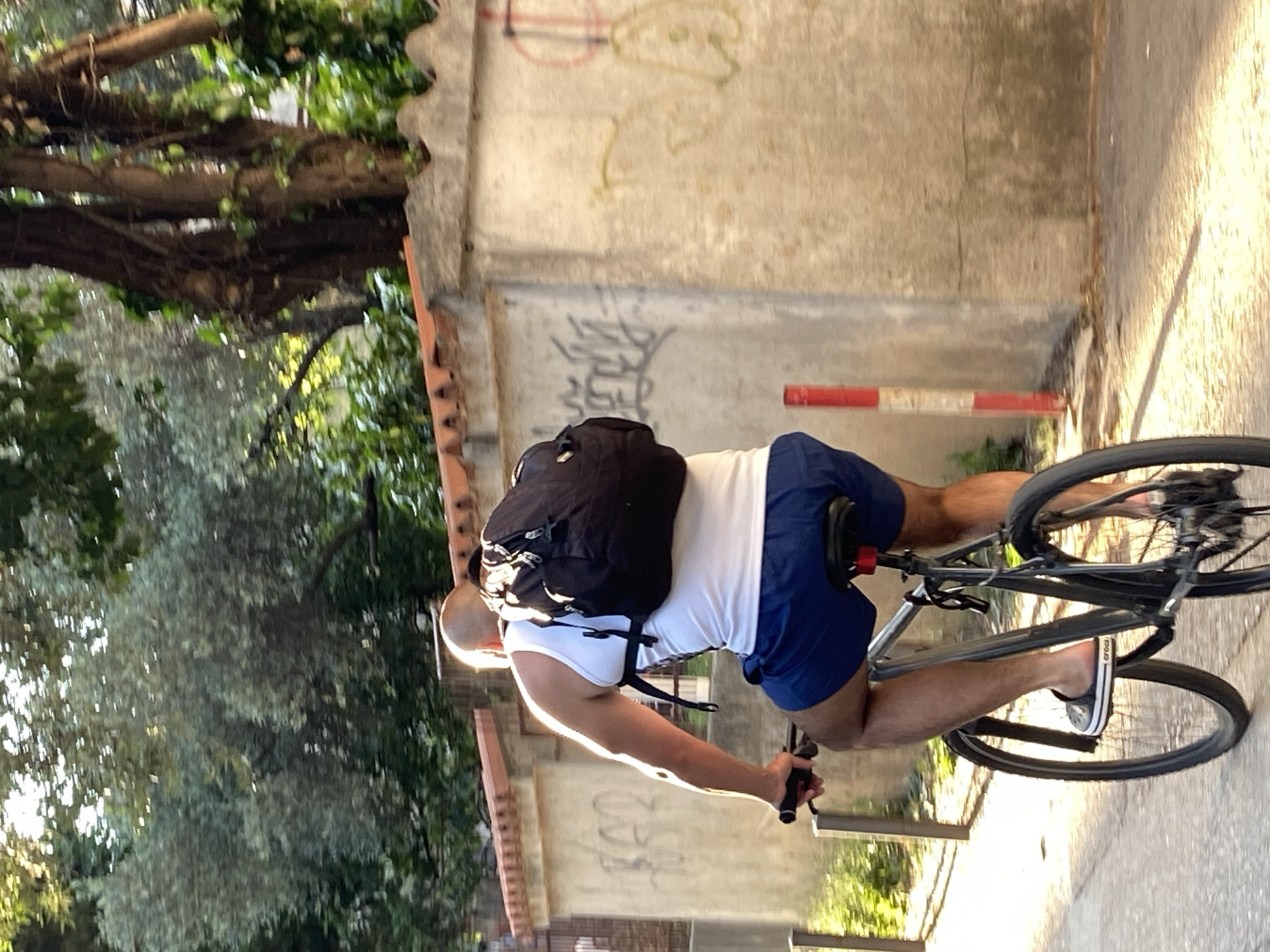 A man rides a bicycle on a path adjacent to a weathered wall, likely in or near the city of Budva, Montenegro, given the architectural style and foliage. The man is wearing a white tank top, blue shorts, and slides. He has a black backpack on his back. The bicycle is a silver-colored road bike. The path is paved and bordered by trees and foliage on one side, with a wall on the other side. The wall has some graffiti on it. A red and white striped pole is visible next to the wall. The lighting suggests the photo was taken in the late afternoon or early morning.