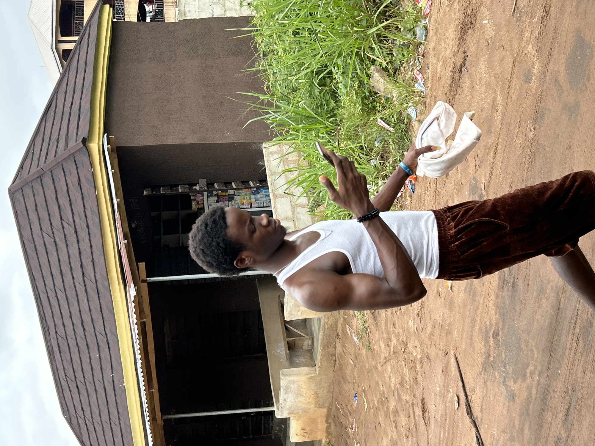 A young Black male is walking on a dirt road in Ihiagwa, Nigeria, looking at his smartphone. He is wearing a white tank top and brown shorts. He is holding a white folded cloth in his right hand. Behind him is a small building with a brown exterior and a dark roof. Lush green grass and some scattered debris are visible to the right of the building. The sky is overcast.