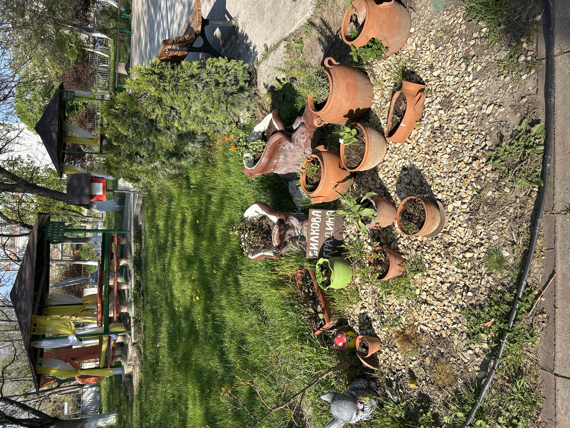 The scene depicts a garden space in Plovdiv, Bulgaria. Dispersed on a gravel surface are various clay pots, some containing plants, others empty or broken. A sign with Cyrillic script, possibly denoting the garden's name, stands amidst green grass. A small, painted ceramic mushroom and a gray rabbit statue add decorative elements. In the background, a covered pavilion structure with hanging decorative fabrics is visible. To the left, a grassy area extends with additional garden ornaments and plant life. A concrete path borders the garden space. The lighting suggests it is daytime.