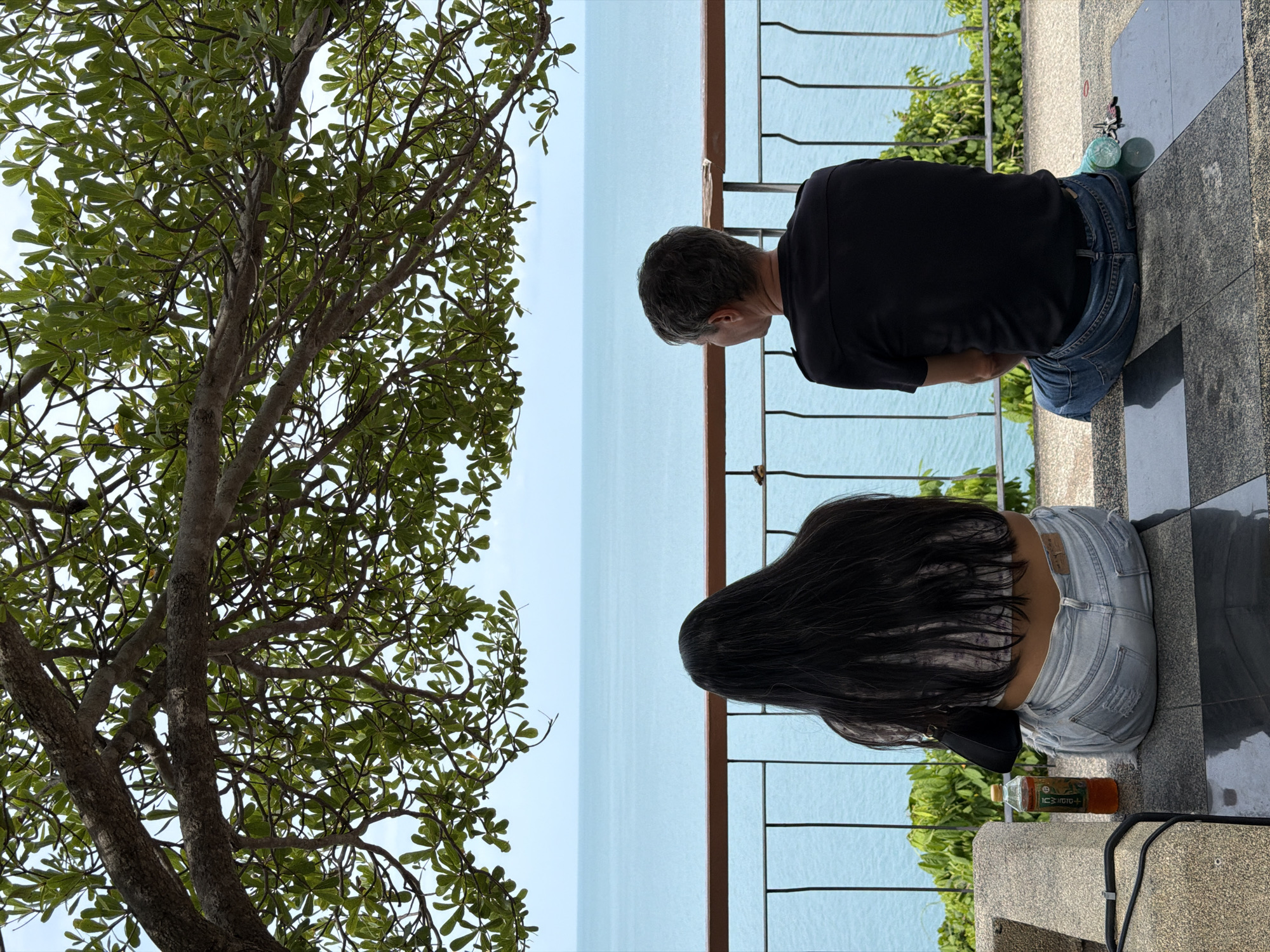 Two individuals, a woman with long dark hair and a man with short grey hair, are seated on a stone ledge, facing away from the camera and towards a body of water. The woman is wearing a patterned top and ripped light-wash jeans. The man is wearing a black t-shirt and blue jeans. A bottle of iced tea and a small black purse are near the woman. A light blue plastic bottle and keys are near the man. They are positioned below a large tree with green leaves, and in front of a railing overlooking the ocean. The ocean is a calm, light blue, meeting a pale blue sky. Green foliage is visible on either side of the railing. The setting appears to be an outdoor viewpoint in the Ko Samui District of Thailand, offering a serene view of the sea.