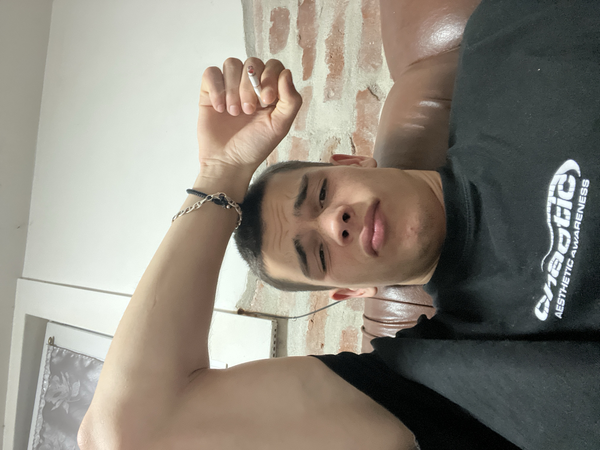 A young man is pictured from a low-angle perspective, seemingly lying down. He is holding a lit cigarette between his fingers on his right hand. He is wearing a black t-shirt and a bracelet. The background includes a white wall on the left and a brick wall on the upper-right. A leather couch or chair is visible behind him. The location is unknown and cannot be determined from the image, and thus cannot be associated with Plovdiv, Bulgaria.