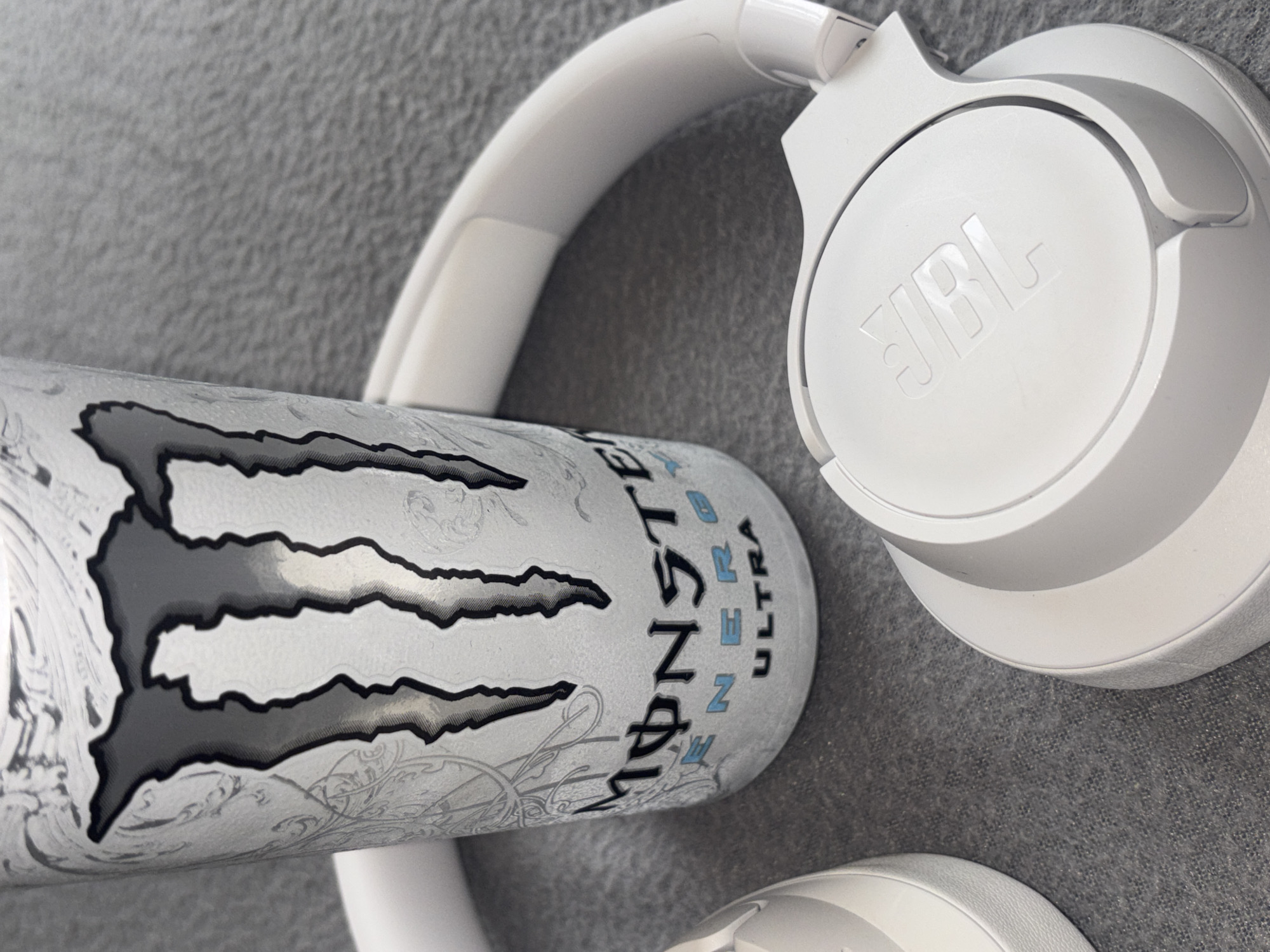 On a textured gray surface rests a white JBL headphone set with the brand logo visible on the right earcup. Beside the headphones is a Monster Energy Ultra can with its distinctive logo. There is no indication of a specific location in Plovdiv, Bulgaria, or any information related to people, actions, or interactions. The image solely shows inanimate objects on a gray background.