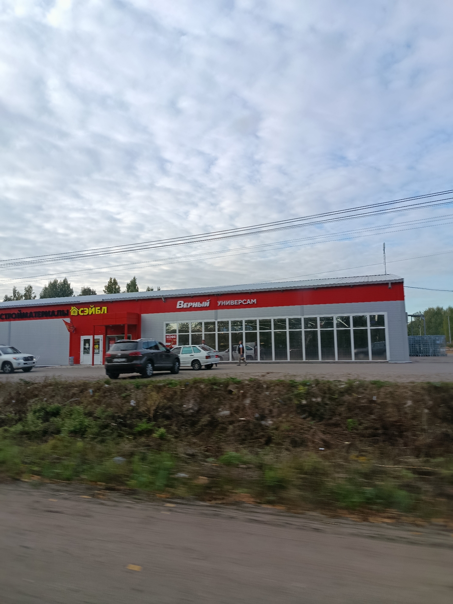 A low-angle shot captures a commercial building with a red and gray facade under a cloudy sky. The building features a row of large windows reflecting the surroundings. A prominent sign above the windows reads "ВЕРНЫЙ УНИВЕРСАМ" in white lettering, with a smaller sign to the left that says "СТРОЙМАТЕРИАЛЫ" above a logo that appears to be a house with "СЭЙБЛ" written next to it. Several cars are parked or driving in front of the building, including a dark-colored SUV and a white sedan. A person is walking on the sidewalk in front of the windows. The foreground shows a blurred embankment covered in dry vegetation and some green growth, suggesting the image was taken from a moving vehicle.