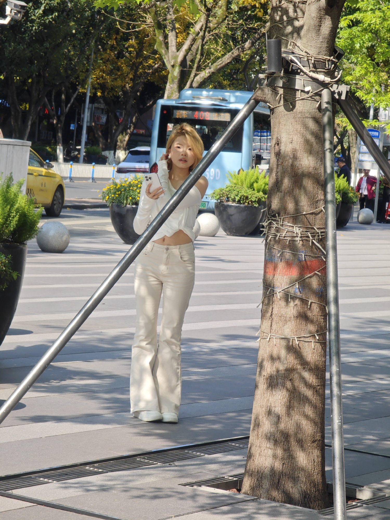 The full-length image captures a young East Asian woman standing on a striped paved urban square in Yuzhong District, China, under bright sunlight. She is positioned slightly to the left of a large tree trunk, which is secured by a prominent metal support structure. The woman wears a long-sleeved, off-the-shoulder white cropped top, light beige wide-leg pants, and white sneakers. She holds a white smartphone in her left hand, positioned near her chest, and looks towards the camera with a neutral expression.
The tree trunk is a central element, adorned with string lights wrapped around it and marked with horizontal red and blue paint bands at mid-height. The metal support structure, composed of black and silver bars, helps stabilize the tree. In the foreground, part of a metal drainage grate is visible on the pavement.
The background reveals a bustling street scene. A light blue public bus with the number "408" and the Chinese character "渝" (Yú, abbreviation for Chongqing) on its front is prominent. Below the number, "S9D" is also visible. A yellow taxi is partially seen on the left, and other vehicles and buildings line the street. Several large, dark planters holding lush green foliage are interspersed around the square. Distinct shadows cast by trees and structures indicate a sunny day. People are faintly visible in the far background.