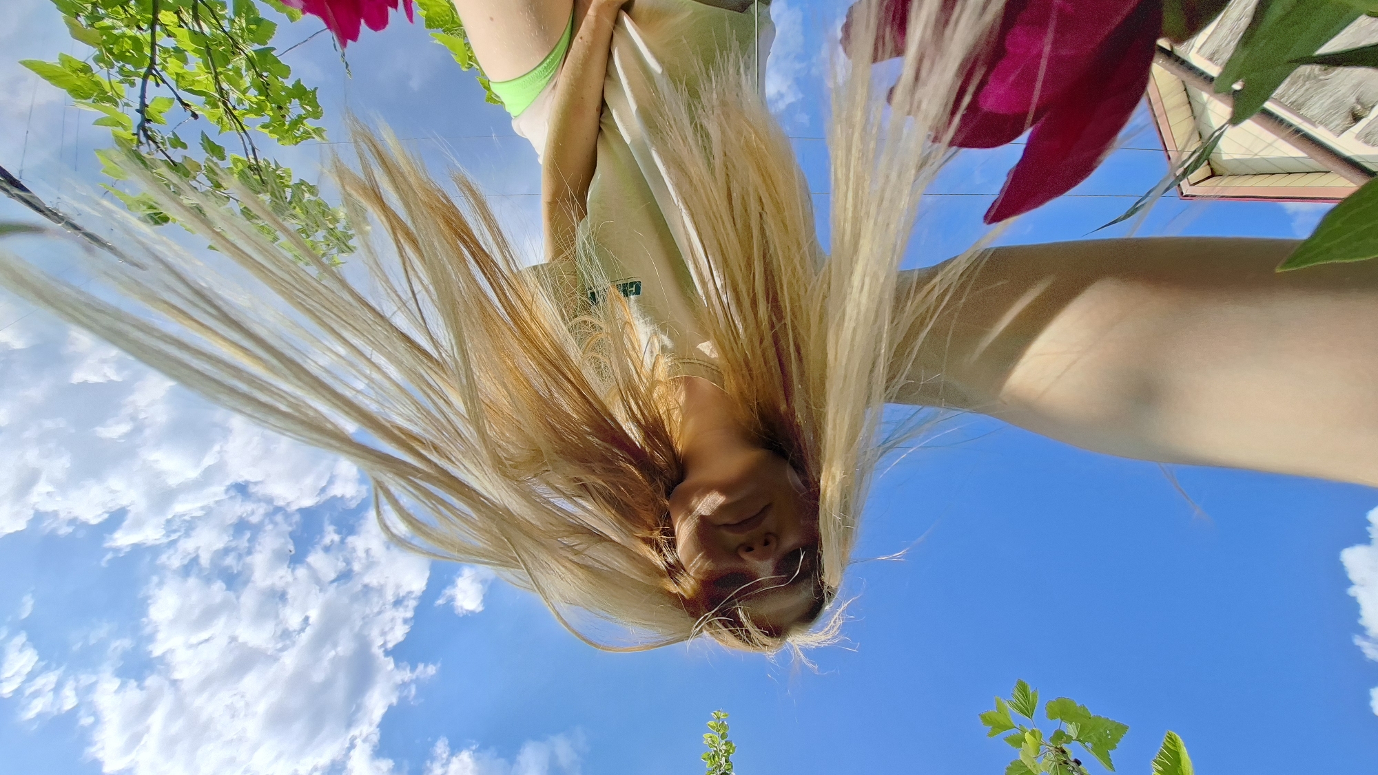 The image is an extreme low-angle, wide-shot selfie taken outdoors on a sunny day with a blue sky and scattered clouds. A young Caucasian female with long, blonde hair is the central subject. She is wearing a light-colored t-shirt and lime green bottoms, and sunglasses. Her hair is blowing in the wind, obscuring parts of her face and the sky. In the foreground, out-of-focus bright pink flowers and green foliage are visible. To the left, the corner of a building with a light-colored facade and a pink-tiled roof is partially visible, suggesting an urban or suburban environment. The overall impression is one of a candid, spontaneous moment captured outdoors, possibly in a residential area of Большой Букор, Russia.