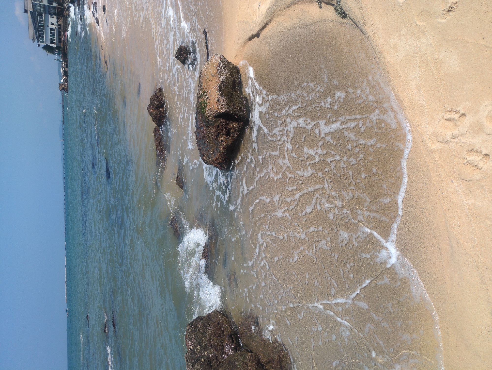 This is an aerial view photograph depicting a sandy beach landscape in Phú Quốc, Vietnam. The composition is vertically oriented, with the waterline and beach running diagonally across the frame. The scene includes coarse sand, several large rocks, and receding seawater. The water is clear enough to see the sandy bottom near the shore. The waves are creating frothy foam as they meet the sand. Two sets of footprints can be seen in the drier sand. In the distance on the left, building construction can be seen. The sky is a light, clear blue.