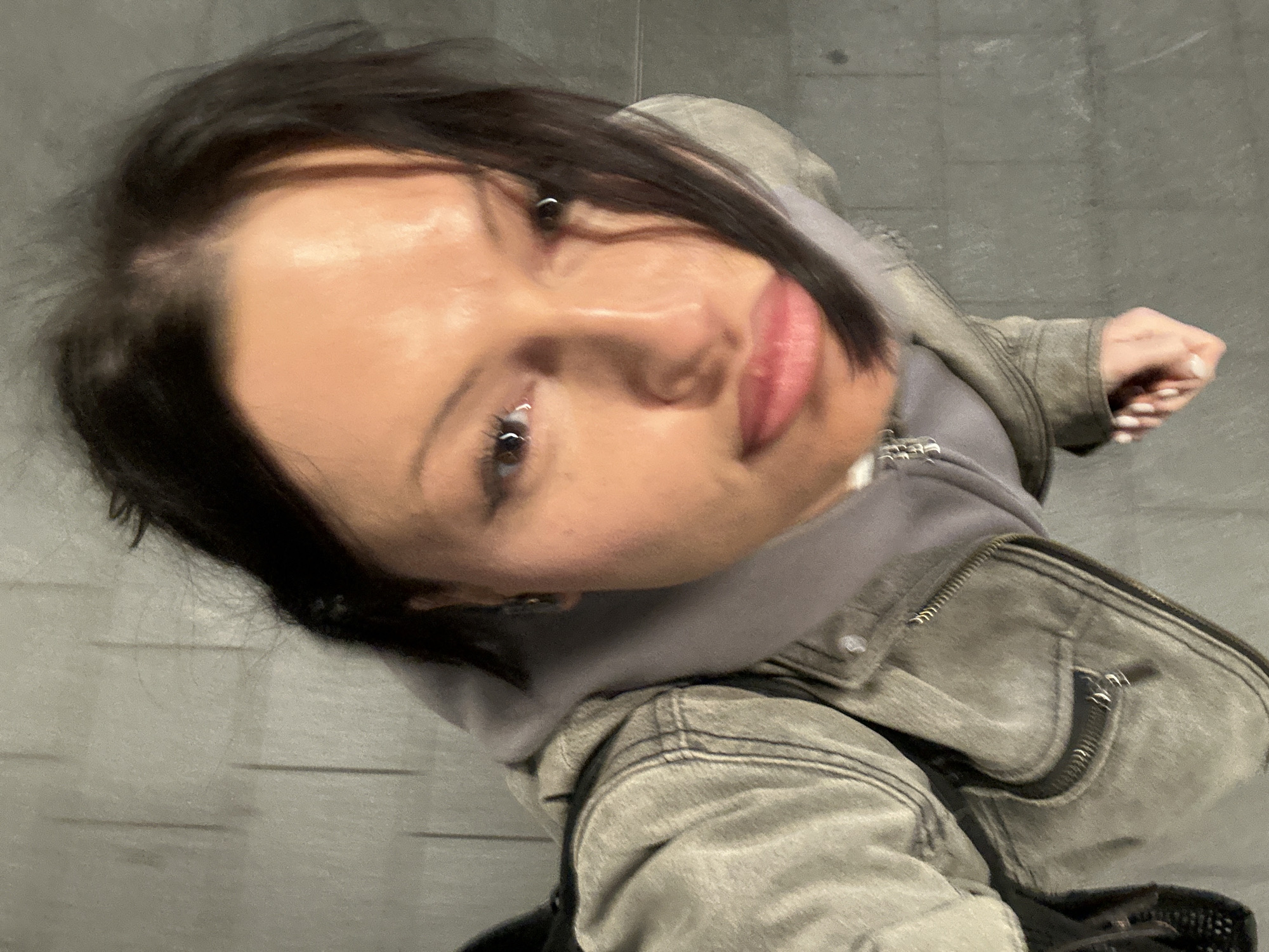 A female individual is depicted in an overhead, slightly tilted portrait, looking directly at the camera. She possesses dark hair, a medium skin tone, and visible facial features include dark eyeliner, a septum piercing, and light pink lips. Attire consists of a light olive-gray denim-style jacket with visible stitching and a zippered pocket on the left, layered over a light gray hooded sweatshirt. Small earrings are present, and her fingernails are painted white. Her right hand is partially visible, appearing loosely fisted. The background is a light gray, textured wall with subtle vertical lines, while the lower right portion of the image displays a darker gray, textured ground surface with linear patterns. The specific location within the city of London, United Kingdom, cannot be determined from the available visual information.