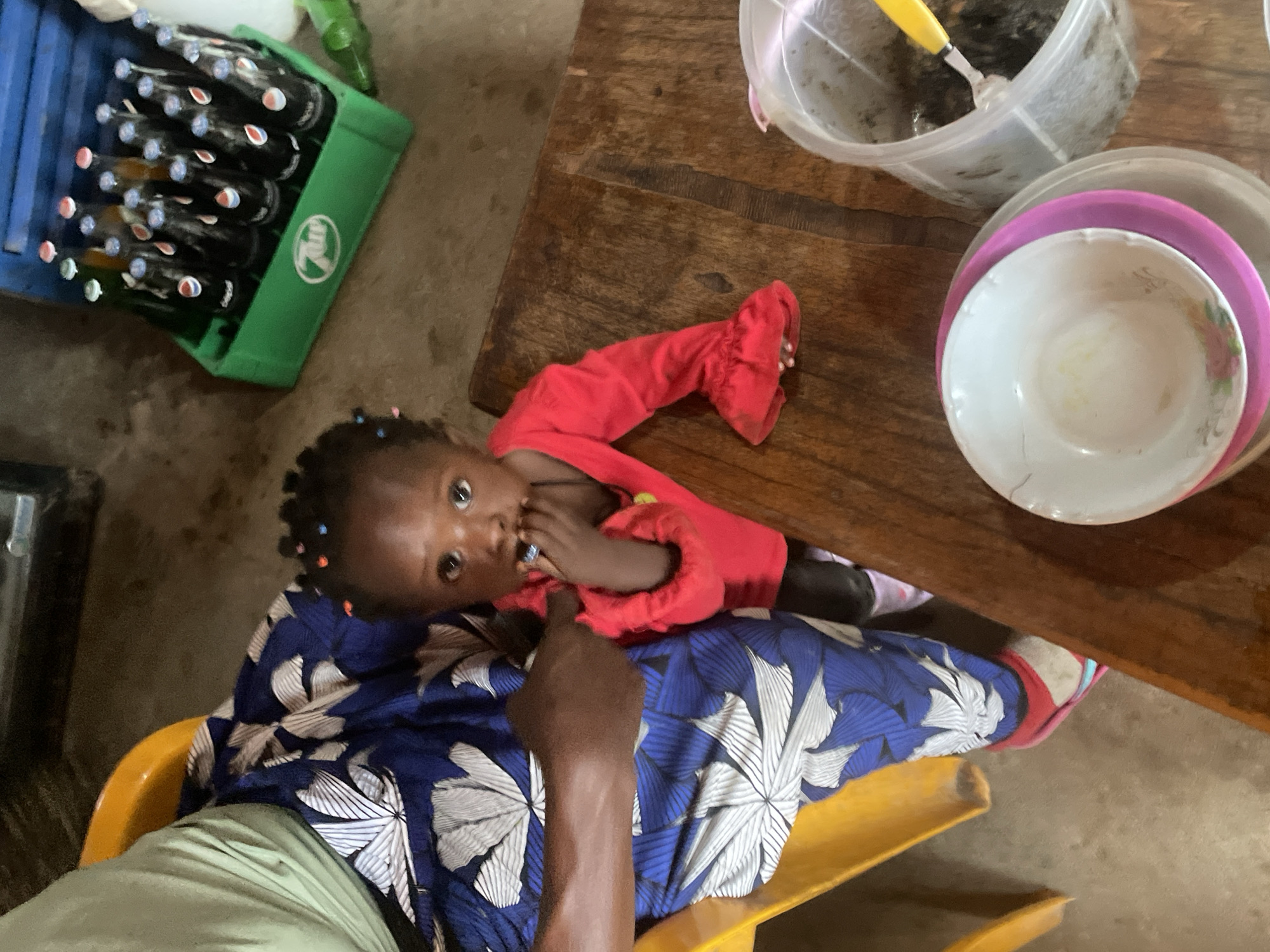 A child in a red outfit with gathered sleeves lies on the lap of an adult wearing a blue and white patterned garment seated in a yellow plastic chair. The child is dark-skinned and has hair adorned with small, colorful beads. The child's hand is near their mouth.
A dark wooden table is beside the child. On the table, there is a stack of white and pink bowls, a plastic container filled with a dark substance and a yellow utensil, and a crate of bottled beverages.
The location is Jos, Nigeria.