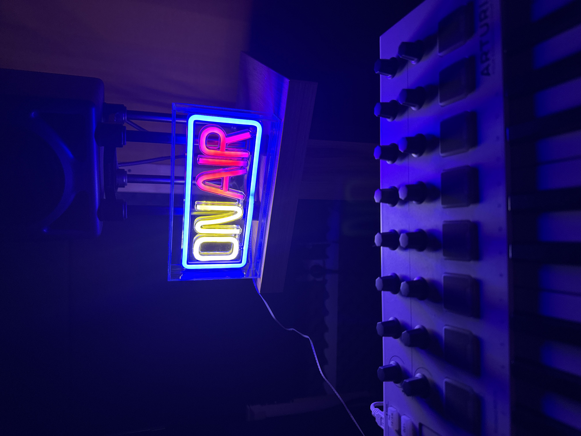 The image shows an "ON AIR" neon sign with a yellow "ON" and a red "AIR," illuminated in a blue-lit room. The sign is mounted on a stand connected to a microphone boom arm. To the right, a white Arturia synthesizer is visible. The location in the city of Plovdiv, Bulgaria, is not identifiable from this image.