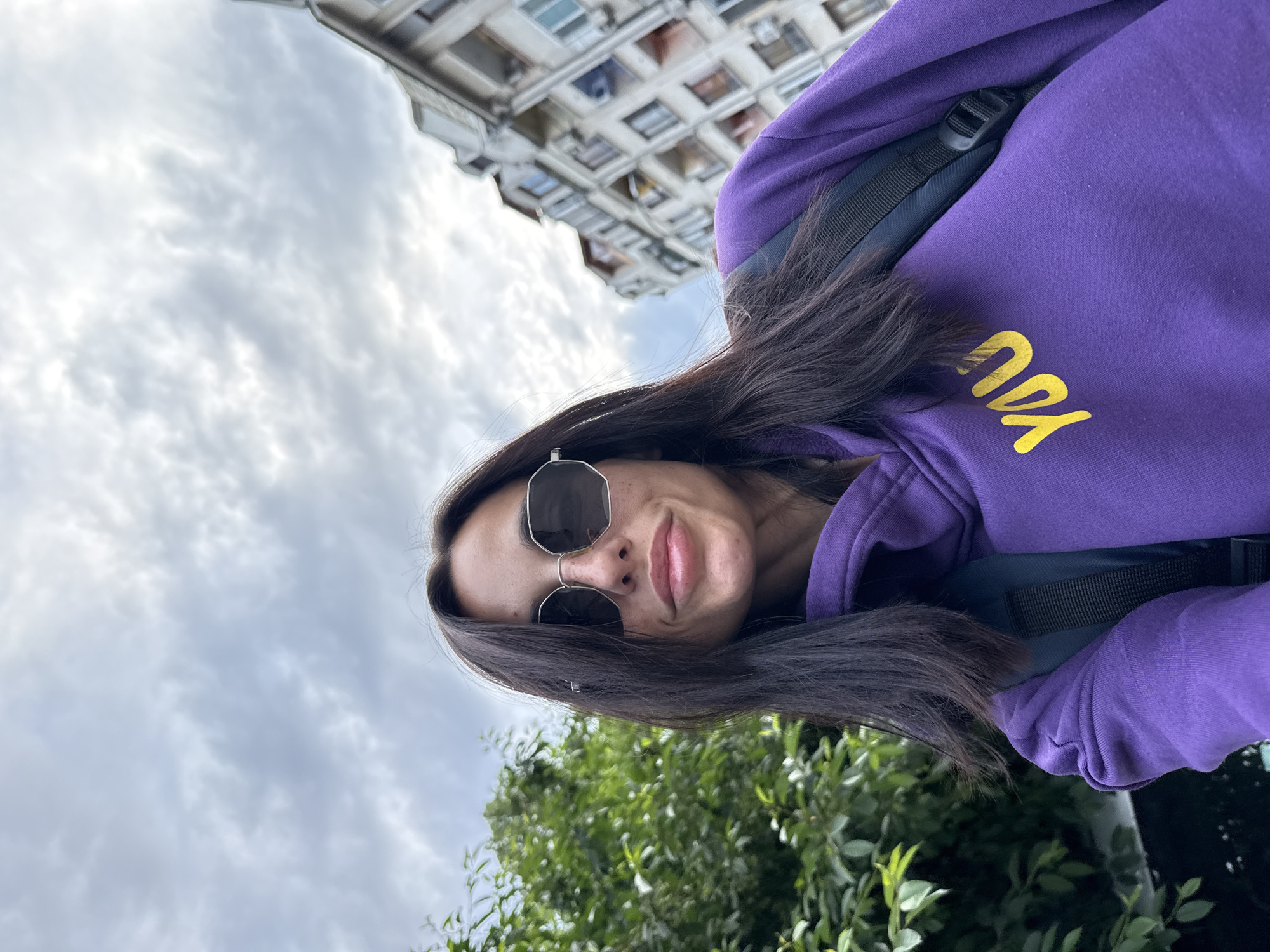 A woman with long, dark hair and wearing sunglasses is pictured in an eye-level, rotated selfie outdoors. She is wearing a purple hoodie with the word "YOU" in yellow on the chest, along with a backpack. In the background, there is a building with multiple windows and a cloudy sky. Green foliage is also visible near the bottom of the image. Although exact city identification is not possible through visual analysis, given the prompt and context that it is in Pleven, Bulgaria, the architectural style suggests a residential building typical of a Balkan city. The location could be near a park or residential area.