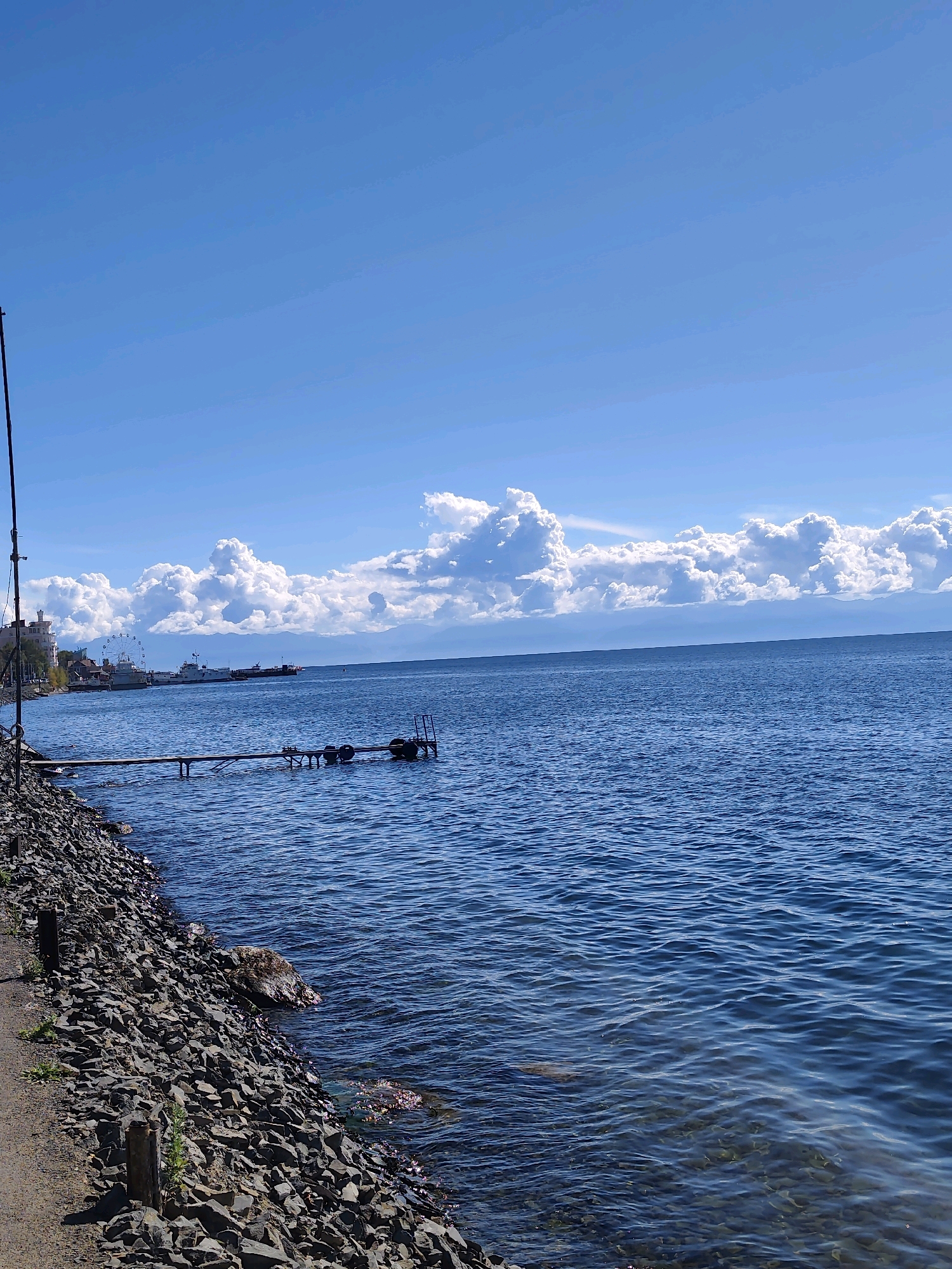 The image captures a view of Lake Baikal from the shore in Listvyanka, Russia. The foreground features a rocky shoreline with a dirt path running alongside it. A tall, thin pole, possibly a fishing rod or antenna, is visible on the far left. The water of the lake is a deep blue, with gentle ripples reflecting the sunlight. A wooden pier extends out into the water, with what appear to be buoys attached. In the middle ground, several boats are docked or anchored along the shore. Behind the boats, there's a glimpse of buildings and a Ferris wheel, suggesting a populated area or amusement park. The sky is a bright blue, with a prominent line of fluffy white cumulus clouds stretching across the horizon. Distant mountains are visible through a haze beneath the clouds. The overall scene depicts a calm day at the lakeside with evidence of human activity.