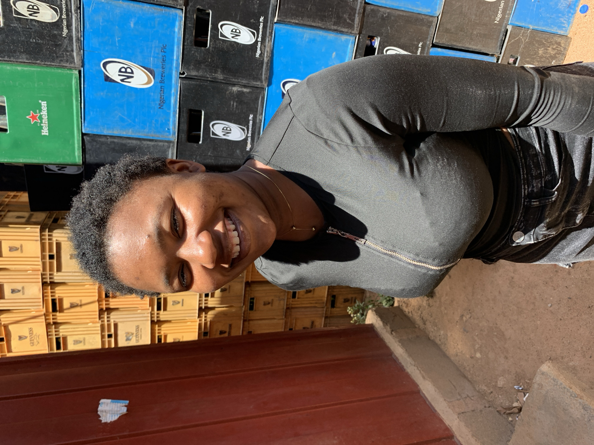 A woman smiles towards the camera while standing next to stacks of beer crates. Some of the crates are branded with "Guinness" and "Heineken" logos. Other crates, stacked higher up, have "NB" (Nigerian Breweries) logos printed on them. The scene appears to be outside, with a concrete step and some dry ground visible in the foreground. The location is in Gura Top, Nigeria.