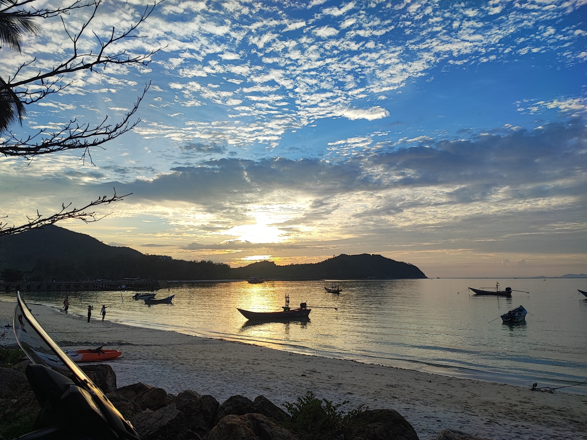 This is a wide-angle, landscape photograph taken at dusk on a beach in the Ko Pha-ngan District, Thailand. The sun is low on the horizon, casting a golden glow across the water and reflecting on the wet sand. The sky is a vibrant blue with scattered white clouds, some of which are tinged with orange from the setting sun.
In the foreground, the beach is made of light-colored sand, with a few large rocks scattered along the shoreline. A dark, reflective object, possibly a boat or a piece of equipment, is positioned in the lower left corner of the frame, partially obscuring the view of the beach. A bright orange kayak lies on the sand to the right of this object. Further down the beach, a few people are visible, appearing to be walking or playing near the water's edge.
The ocean is calm, with gentle waves lapping at the shore. Several small boats are anchored in the water, some of them silhouetted against the bright sky. In the distance, dark, tree-covered hills form the coastline. The overall scene evokes a peaceful and serene atmosphere.