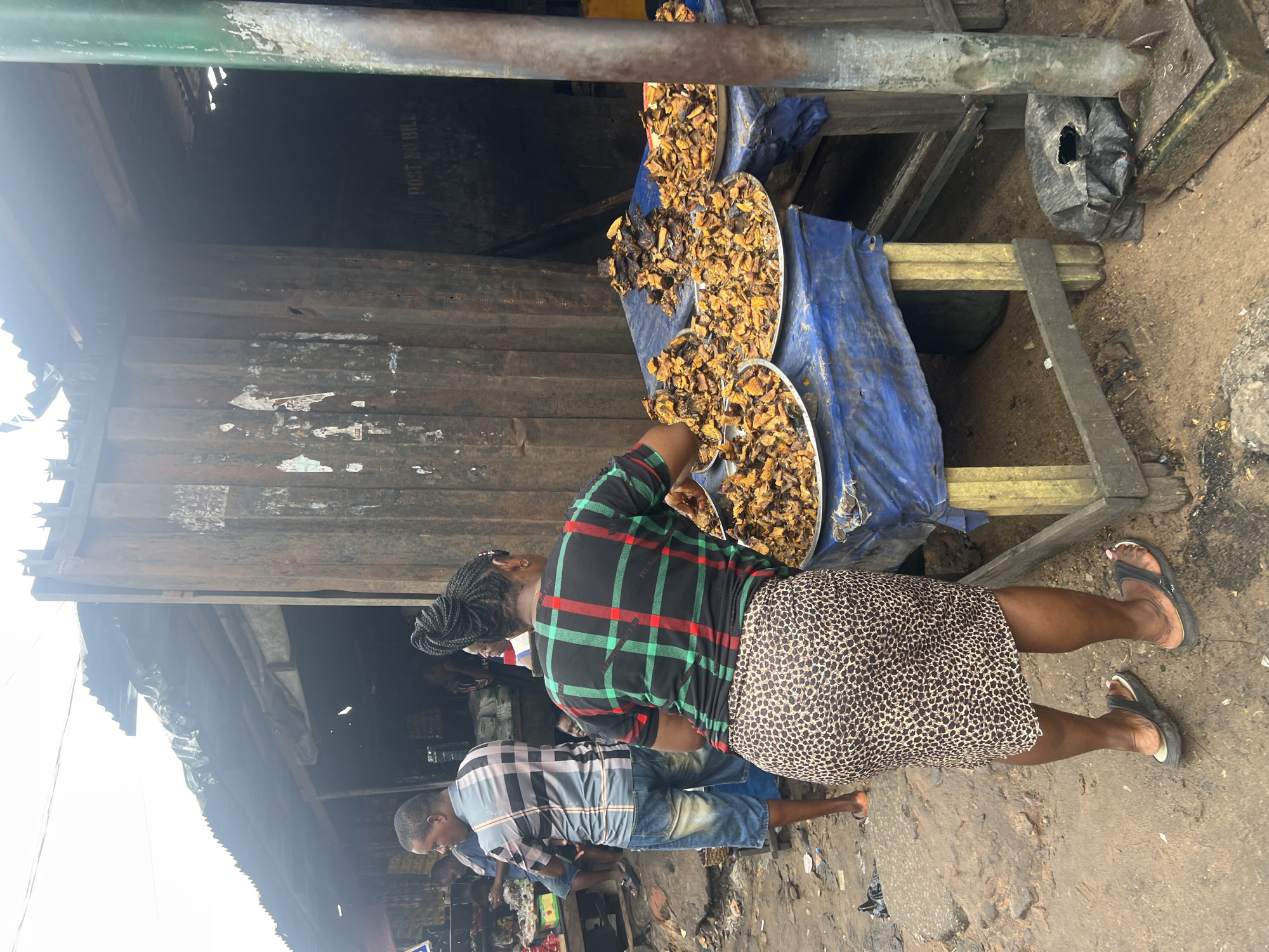 A woman, viewed from behind, stands at a market stall in Nkpor, Nigeria. She is wearing a black, green, and red plaid shirt and a leopard-print skirt, with black sandals on her feet. Her dark hair is styled in a bun. Her hands are engaged with several metal platters filled with what appears to be dried fish or meat. The stall is draped with a blue tarp, and yellow wooden supports are visible. Behind her, a man in a plaid shirt and denim shorts is standing, also facing away from the camera. The ground is muddy and uneven, with some debris scattered around. The background consists of market structures made of corrugated metal, with some posters and graffiti on the walls. The overall atmosphere suggests a busy, outdoor market environment.