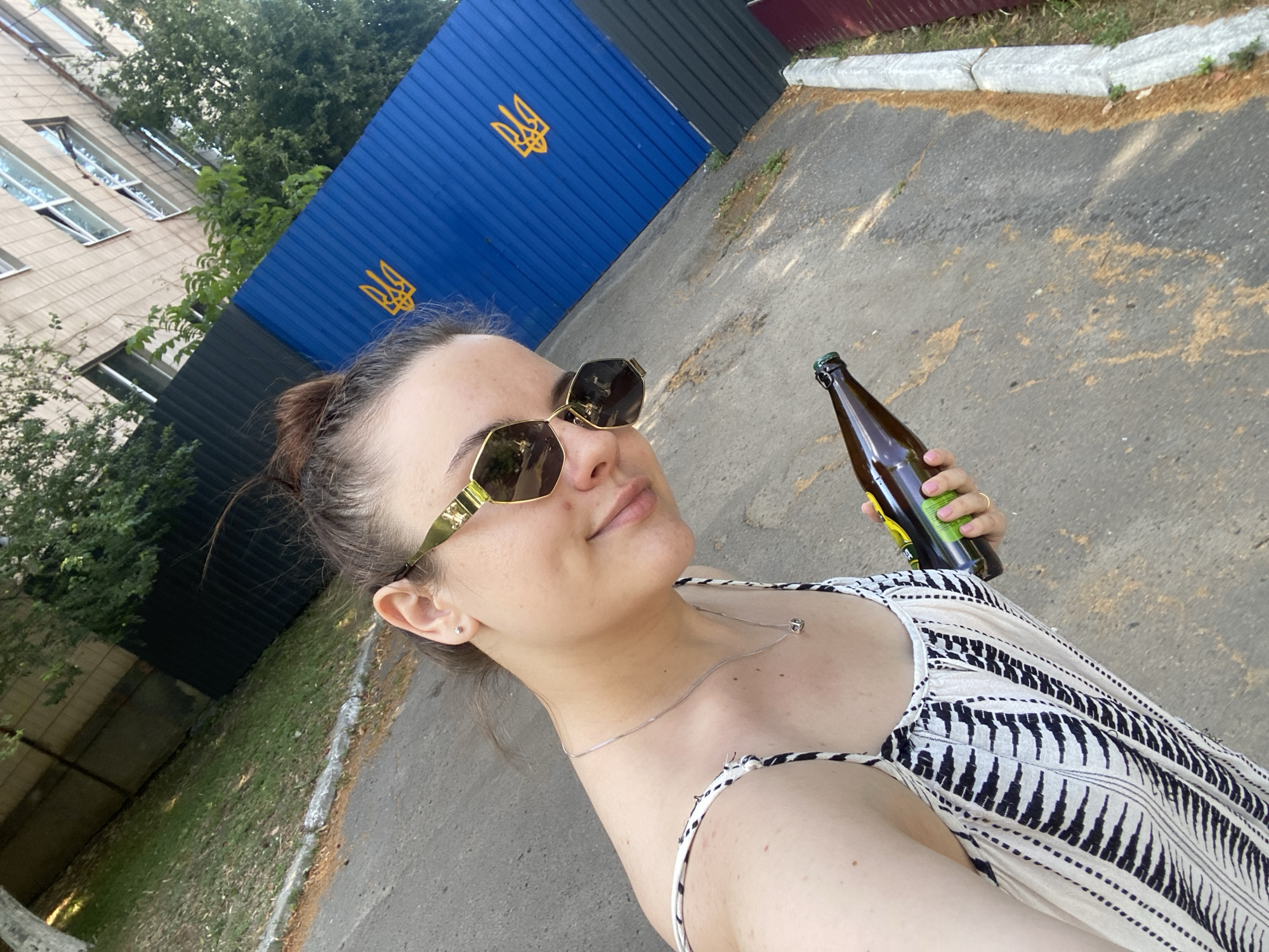 A woman is taking a selfie outdoors in Poltava, Ukraine. She is wearing sunglasses and a patterned dress, and holding a brown glass bottle with a green cap. In the background, there is a blue corrugated metal fence with the Ukrainian trident symbol, a building, trees, and a grassy area. The ground is paved.