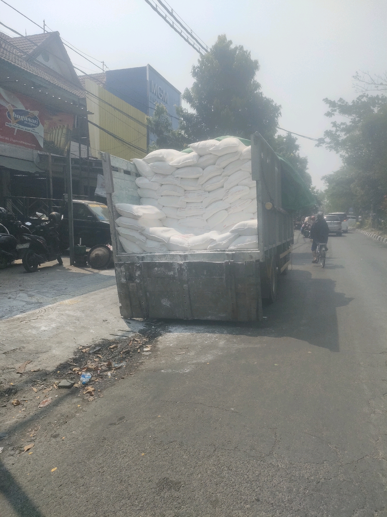 The image shows a truck loaded with large white sacks, likely containing bulk material such as flour or rice, parked on the side of a street. The truck bed is filled to capacity with these sacks, some of which appear to be spilling slightly onto the ground. The truck is situated in what appears to be an urban environment, with buildings visible on the left side of the frame. One building has a sign with "MGM" in large blue letters. Motorcycles are parked on the sidewalk to the left of the truck. The street is paved and appears to be in moderate use, with a few vehicles and a person on a bicycle visible in the distance. The overall lighting suggests it is daytime, possibly with hazy or overcast conditions given the bright, diffused light. The scene suggests a commercial or industrial area, possibly in Surabaya, Indonesia, given the context provided. There is some debris and scattered material on the pavement near the truck.