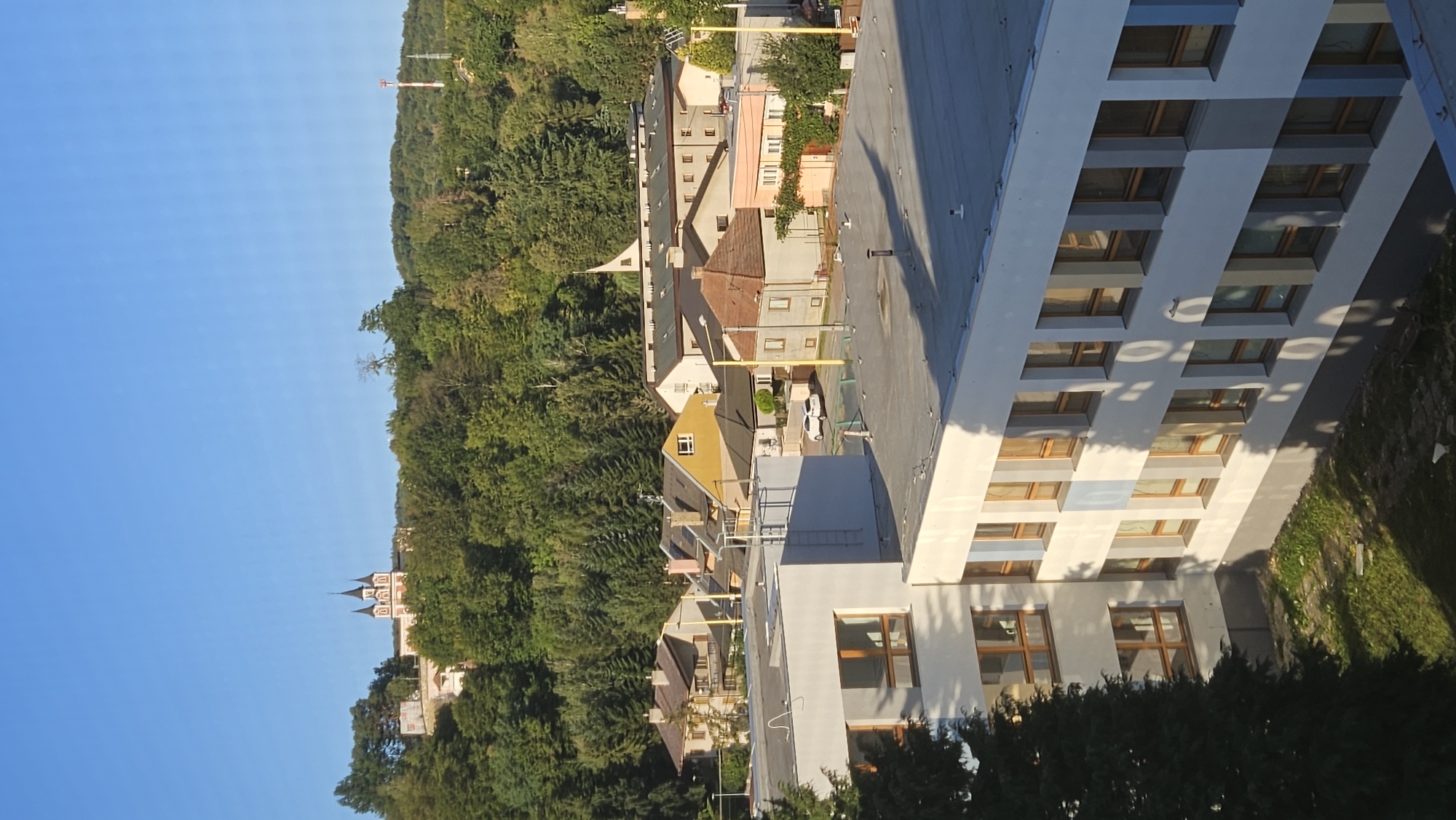 This is a high-angle, vertical shot of a city landscape with a clear blue sky. In the foreground, the upper floors of a light-colored, modern building with multiple windows are visible. The building has a flat, gray roof. In the middle ground, there is a collection of residential buildings with pitched roofs, varying in color and architectural style, with some trees interspersed. Further in the background, a densely wooded hill rises, topped by a pink and white building with dark spires, possibly a church or monastery. A red and white telecommunications tower is also visible on the right side of the hill. The scene appears to be from the city of Prešov, Slovakia, likely overlooking a neighborhood and the surrounding natural environment. The lighting suggests it is daytime, possibly morning or late afternoon due to the long shadows.