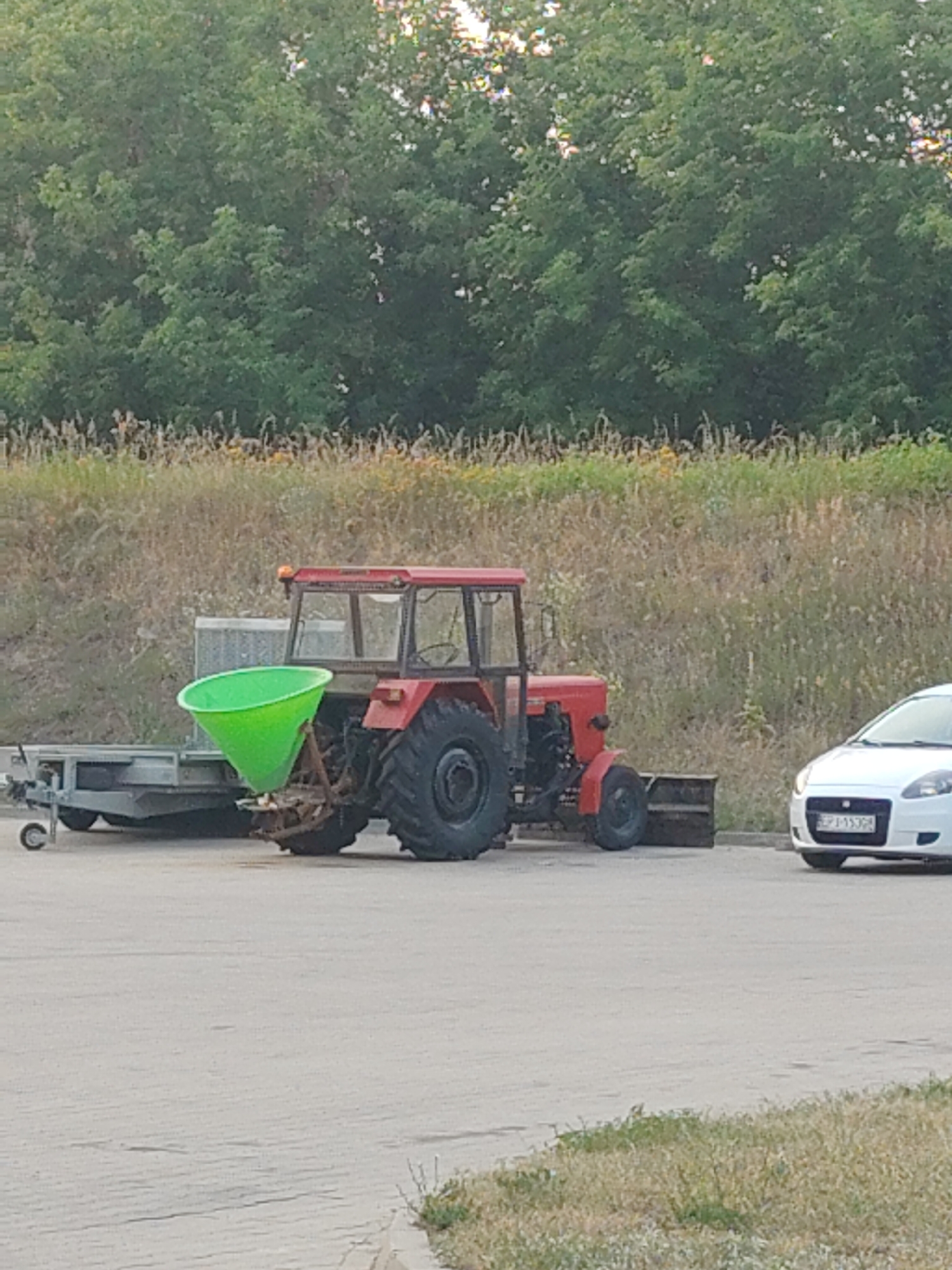 A red tractor with a green hopper attachment is parked on an asphalt surface. The tractor has large rear wheels and a cab. To the left of the tractor is a flatbed trailer. In the background, there is a grassy slope with trees at the top. A white car is parked to the right of the tractor. The scene appears to be outdoors, possibly in a rural or semi-rural area. Based on the car's license plate, which reads "EPJ 1530R," the location is identified as Nowy Dwór Mazowiecki, Poland.