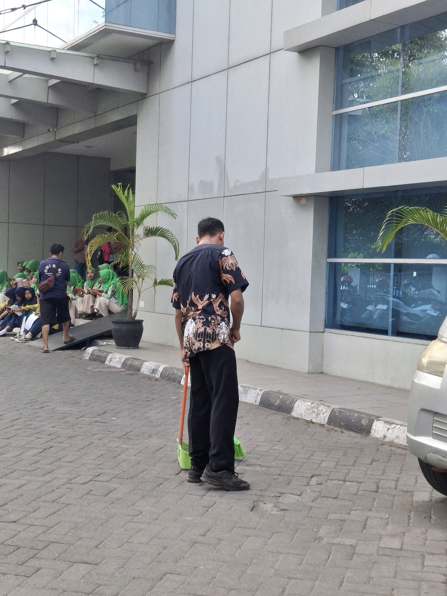 A man wearing a black shirt with an ornate pattern and black pants is standing on a paved area, holding a broom with a green dustpan. He is facing away from the camera. In the background, several young people, mostly female and wearing green headscarves, are sitting on steps in front of a modern building with a glass facade. A palm tree in a pot is situated next to the building. The building appears to be a public or institutional structure. The scene is likely located in Lumajang, Indonesia, given the clothing and general environment.