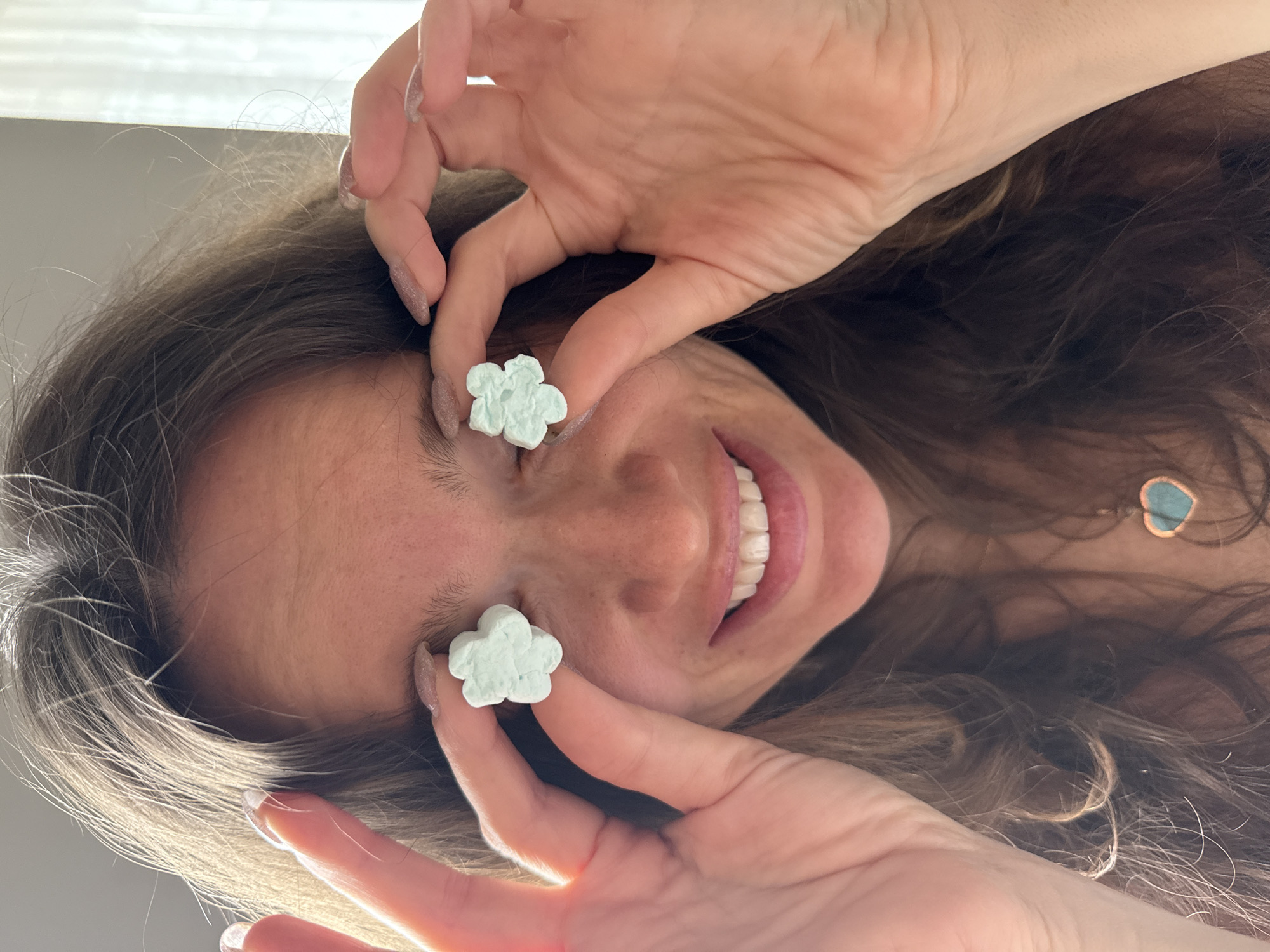 A woman is holding two flower-shaped objects in front of her eyes. The objects appear to be a light blue color. The woman is smiling, and her teeth are visible. She has long, wavy brown hair. A necklace with a heart-shaped pendant is visible around her neck. The image is taken from a low angle, looking up at the woman. There is no indication of the location being Khimki, Russia or any other location in the image.