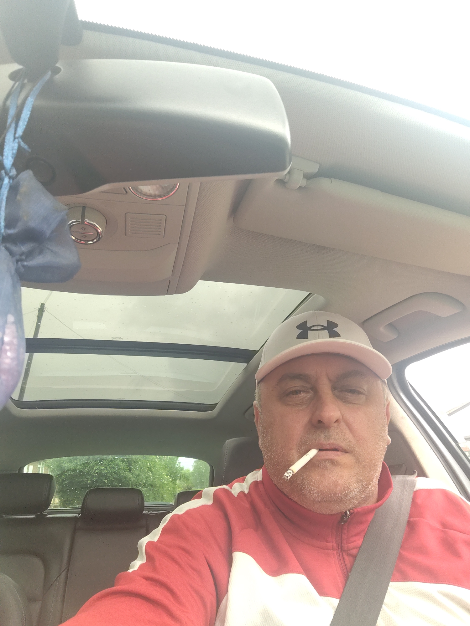 An adult male is captured in a selfie-style shot from the interior of a car. He appears to be seated in the driver's seat, wearing a seatbelt that stretches from his right shoulder across his chest. He has a light-colored (likely white or light grey) baseball cap with a dark, stylized "UA" (Under Armour) logo on the front. He sports stubble or a short beard and has a neutral or slightly tired expression, with a lit or unlit cigarette held between his lips.
He is wearing a red and white track jacket with a zipper down the front. The car's interior is visible, featuring a light grey/beige headliner and trim. An overhead console with a circular dial (possibly for climate control or lighting) and air vents is present. A rear-view mirror with a blue fabric ornament hanging from it is visible in the upper left. A panoramic sunroof extends above, with raindrops clearly visible on its glass, suggesting recent or ongoing rain. Through the rear window, dark-colored back seats are seen, and beyond them, green trees and foliage are visible, indicating an outdoor, possibly suburban or rural, setting. The time of day appears to be overcast daylight.