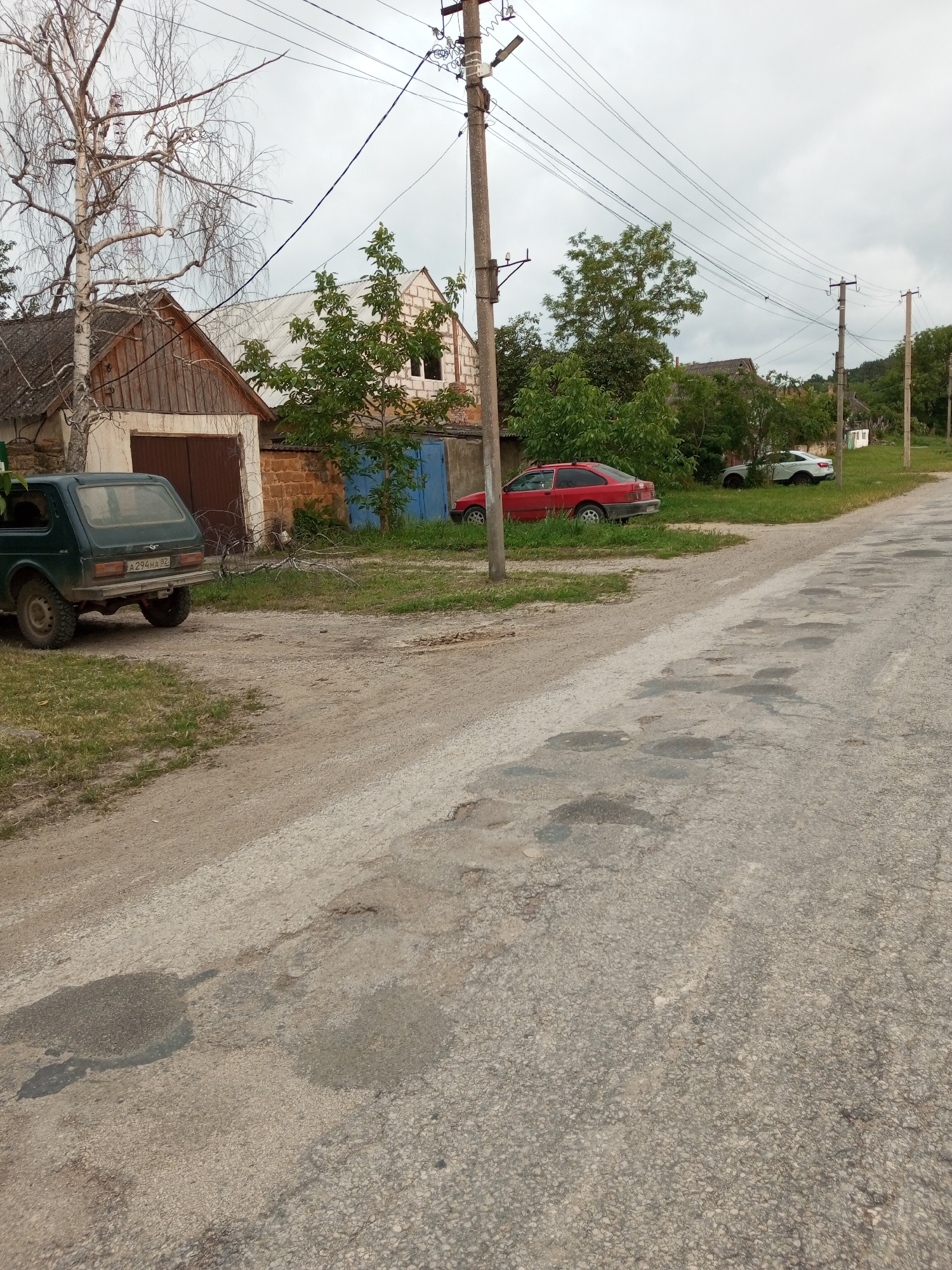 The image depicts a street scene in Ароматновское сельское поселение, Ukraine. The road in the foreground is paved and shows significant signs of disrepair, with numerous potholes and patched areas. To the left of the road, a green SUV is parked, and behind it are two garages. One garage has a wooden door, and the other has a blue metal door. A partially visible brick wall separates these structures. Further back, several trees and buildings are visible. A red sedan is parked on the grass to the right of the road, and behind it, a white SUV is also parked on the grass. Telephone poles and overhead wires run along the street. The sky is overcast.
