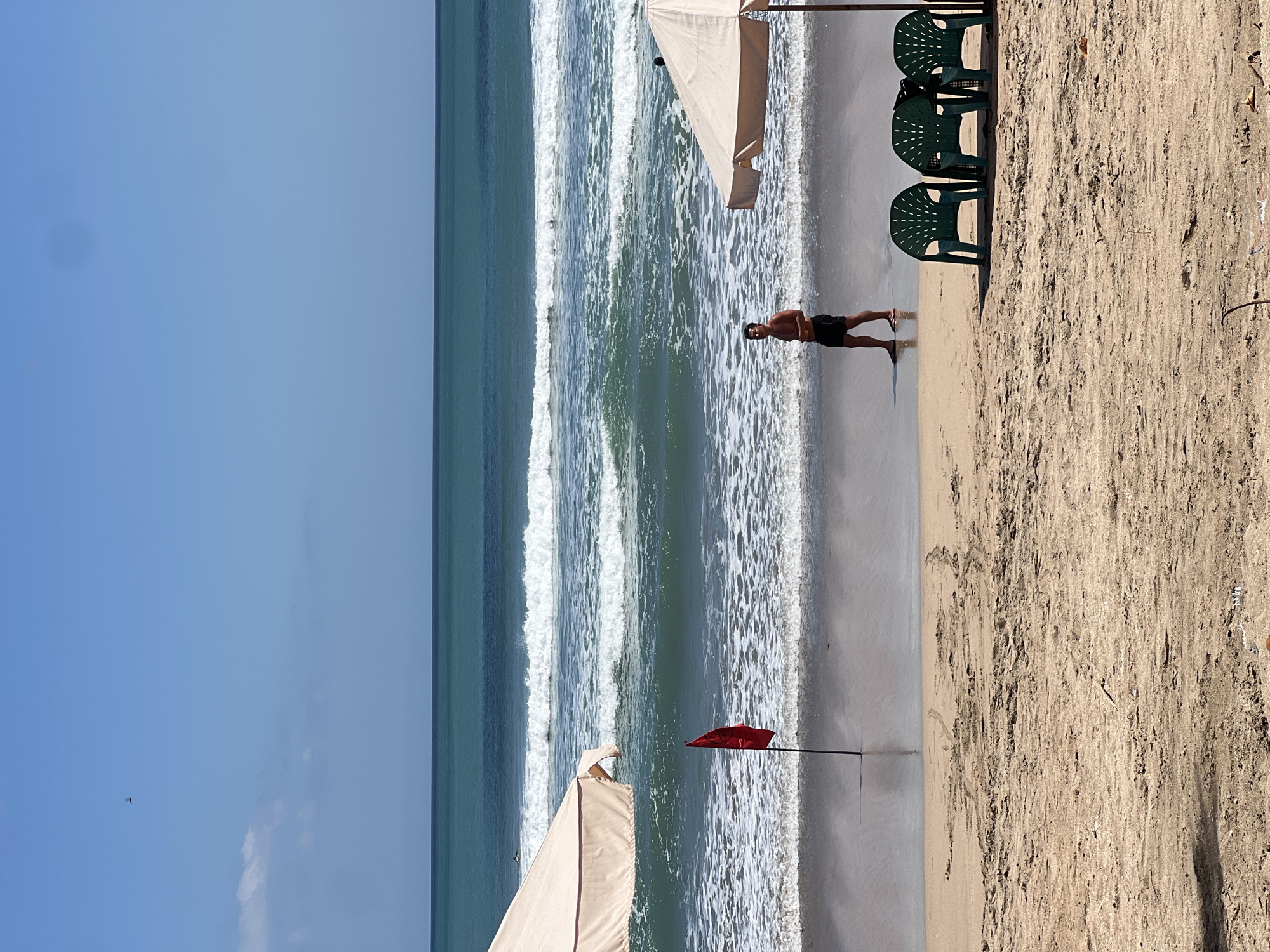 A wide shot captures a serene beach scene in Kuta, Indonesia, under a clear blue sky with a few wispy clouds. The turquoise ocean stretches to the horizon, with gentle waves rolling onto the shore. In the foreground, the sandy beach is mostly empty, with the exception of a few scattered objects. A red flag on a pole stands near the water's edge, signaling a warning. To the right, two beige beach umbrellas provide shade over a group of three green plastic chairs. A lone man, shirtless and wearing black shorts, walks along the wet sand near the water. The overall impression is one of a peaceful and idyllic beach day.