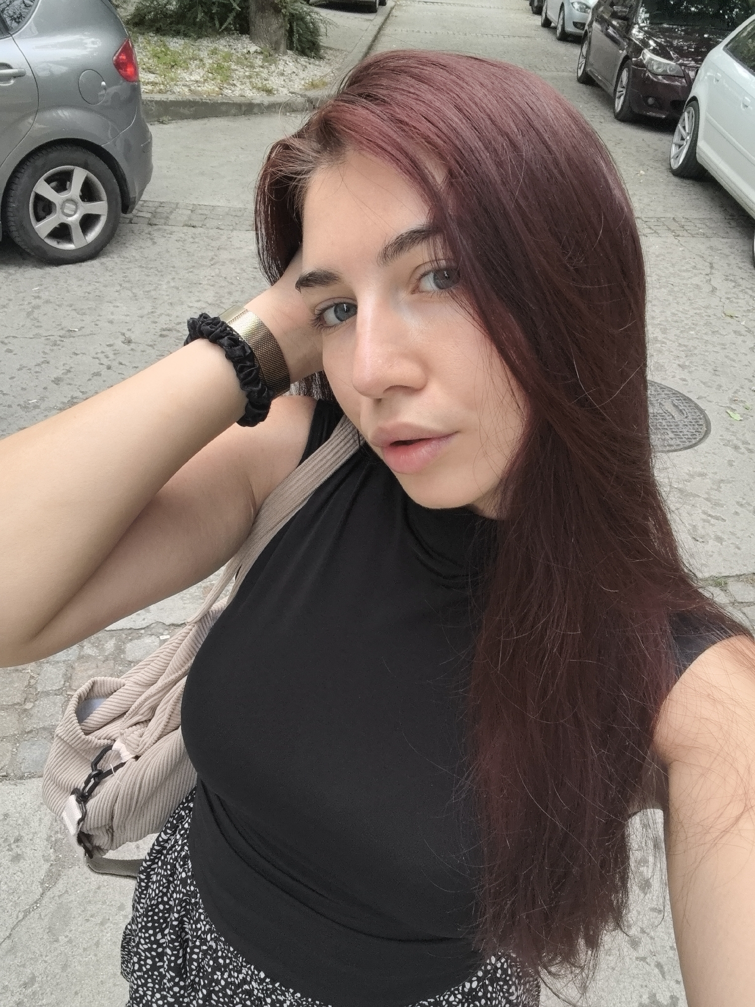 The image shows a young woman standing on a paved street in Stara Zagora, Bulgaria. She has long, dark auburn hair and is wearing a black, sleeveless top and a black and white patterned skirt. A tan corduroy bag is slung over her shoulder. She is holding her left arm up, resting her hand on the back of her head, and is wearing a gold mesh bracelet on her wrist. In the background, several cars are parked along the street, including a silver hatchback on the left and other vehicles further down the road. The ground appears to be asphalt or concrete, with some visible cracks and a manhole cover. The overall scene suggests an outdoor urban environment.