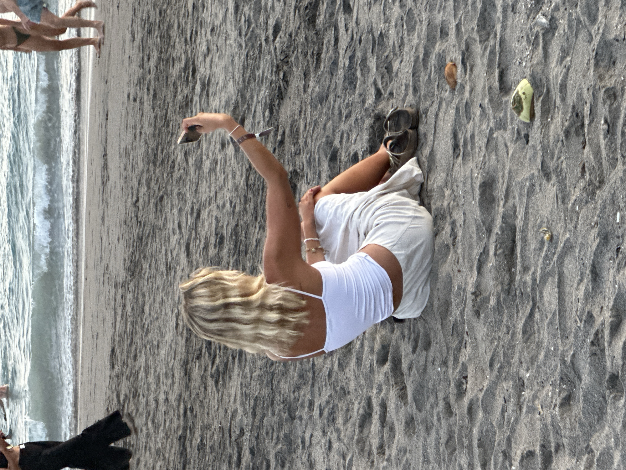 A woman is sitting on a gray sandy beach on the shoreline of the ocean. She is wearing a white tank top and a pair of white shorts or a skirt. She has wavy blonde hair. She has brown sandals beside her. She has an arm raised and appears to be holding a phone. There are people in the ocean in the upper left corner of the image. This photo was likely taken in North Kuta, Indonesia.