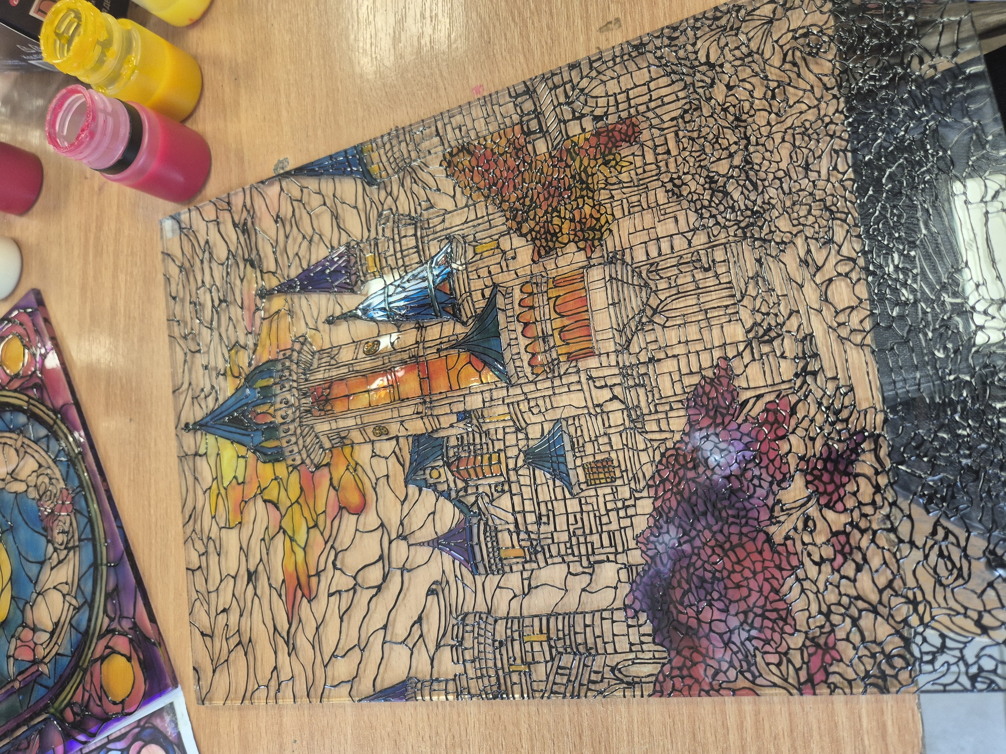 The image shows a handcrafted glass artwork on a wooden surface. The artwork features a futuristic cityscape with rocket ships and abstract forms. The color palette includes yellows, oranges, reds, blues, and purples. The artwork is outlined with a black, crackle-effect border. There are several bottles of colored paint to the side. The artwork resembles stained glass but is created with paint and outline. There is no evidence of the location being in Pavlikeni, Bulgaria.
