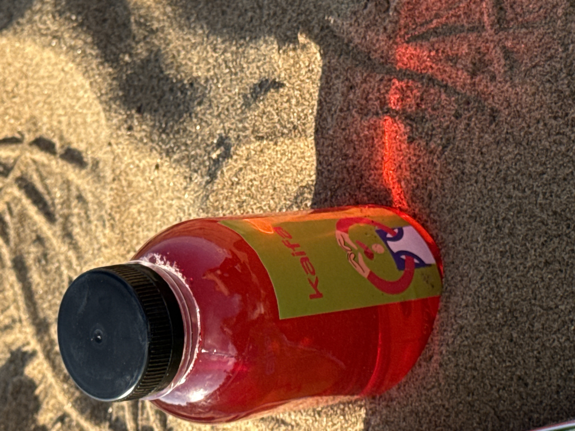 A close-up, high-angle shot features a bright red liquid in a clear plastic bottle with a black cap. The bottle has a green label with red lettering that reads "Kaifa" and a cartoon image. The bottle is placed on a sandy surface, possibly a beach, with shadows cast by the bottle and sunlight. The sand appears granular and is illuminated by warm light, creating a textured background. The overall impression is one of a refreshing drink on a sunny day. The provided image does not contain any information that can be used to determine its location within the city of Chaikovsky, Russia.