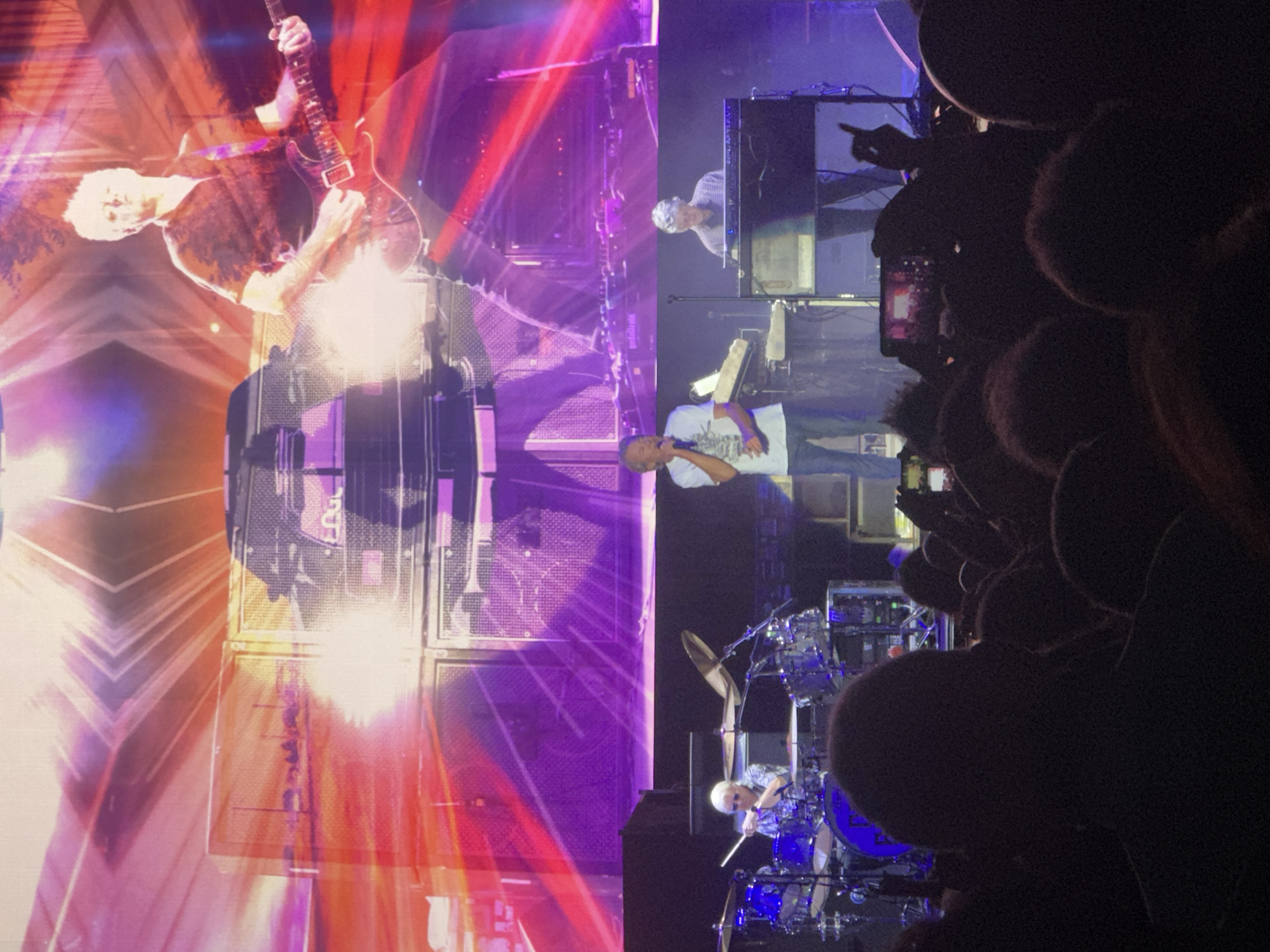 The image captures a live music performance at an indoor venue, likely a concert hall or arena. The stage is dominated by a large screen displaying a vibrant, abstract background with geometric patterns and bright lights in shades of purple, pink, and orange. Several musicians are visible on stage. In the foreground, the audience is partially visible, silhouetted against the stage lights.
Specifically, a guitarist in a dark shirt is playing a red electric guitar. To the left, a drummer with white hair is seated behind a drum kit. In the mid-ground, a singer in a white t-shirt and blue jeans is holding a microphone and appears to be singing. To the right, another musician is playing a keyboard. Various musical instruments and equipment, including amplifiers and keyboards, are arranged on the stage. The atmosphere is energetic, indicated by the lighting and the presence of performers and an audience.
While the image depicts a concert, there is no visual information within the image to suggest that the location is the city of Jerez De La Frontera, Spain.