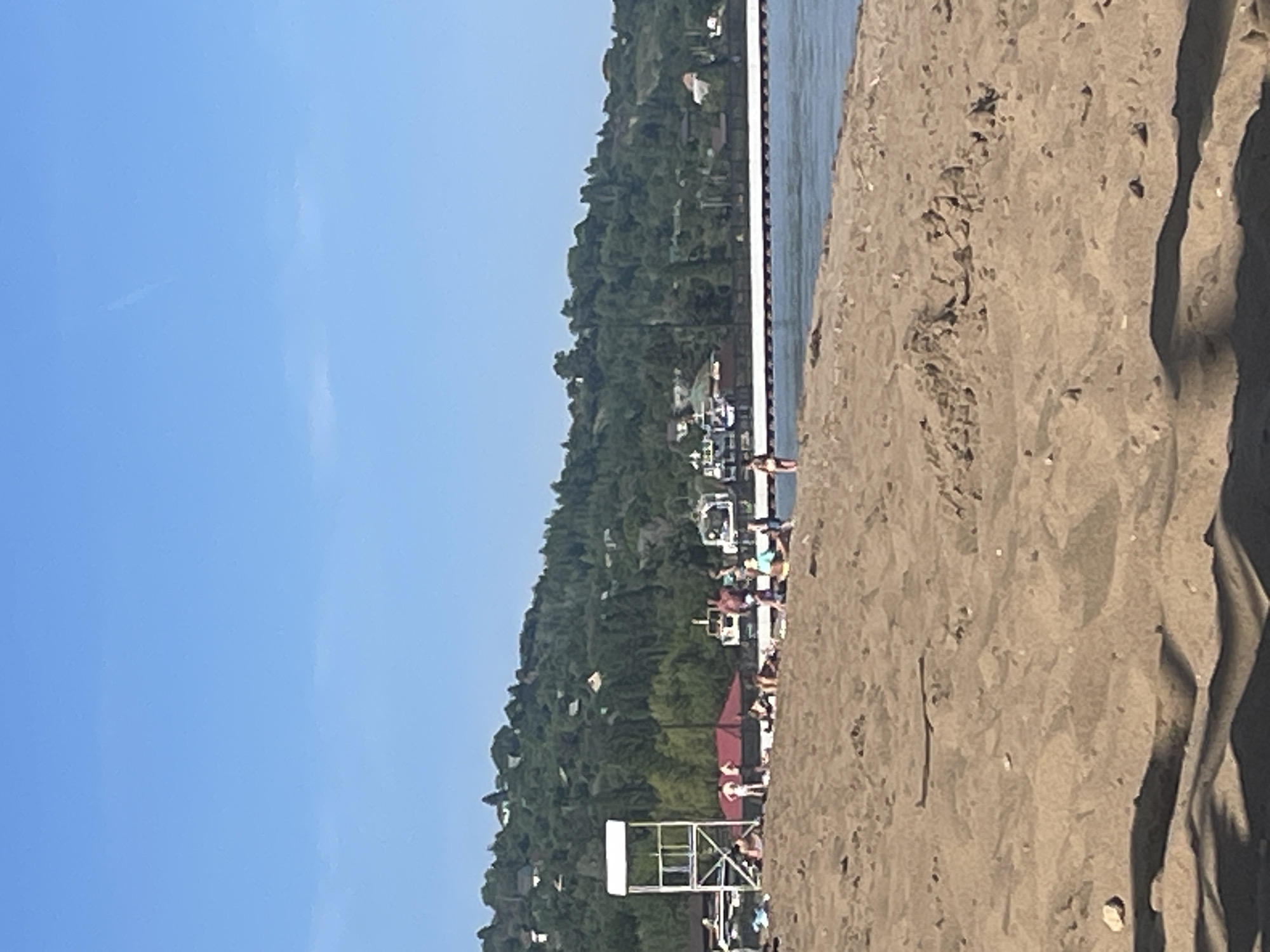 The image shows a beach scene in Saratov, Russia. The foreground consists of a sandy beach with visible footprints and shadows. In the middle ground, there is a lifeguard stand, a group of people on the beach, and a long pier extending into the water. Beyond the pier, a densely wooded hill rises, with scattered buildings visible among the trees. The sky is clear and blue. The image is oriented vertically, with the blue sky dominating the left side and the beach stretching across the right.