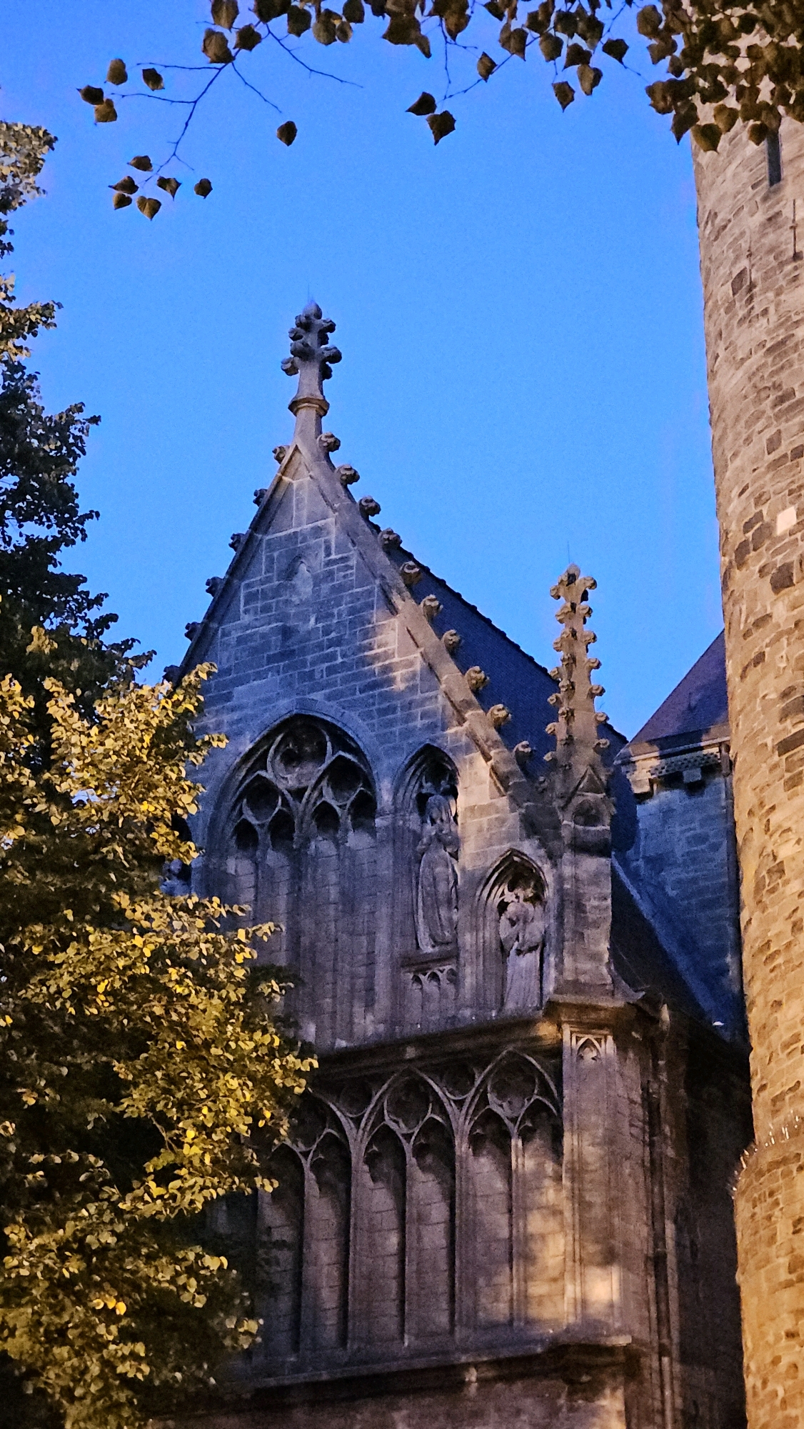 The image shows the exterior of a Gothic-style building, likely a church, under a clear blue twilight sky. The architecture features pointed arches, ornate stonework, and statues carved into niches. Foliage from a tree, with yellow and green leaves, partially obscures the left side of the building. To the right, a portion of a circular brick tower is visible. The lighting suggests it is early evening, with artificial light highlighting some parts of the building. This building is likely part of the Sint Servaasbasiliek in Maastricht, Netherlands, given its architectural style and prominence in the city. There are no people visible in the image.